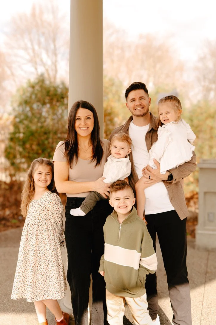 Family of six posing outdoors in fall, with trees in the background. Two adults and four children are smiling and looking at the camera.