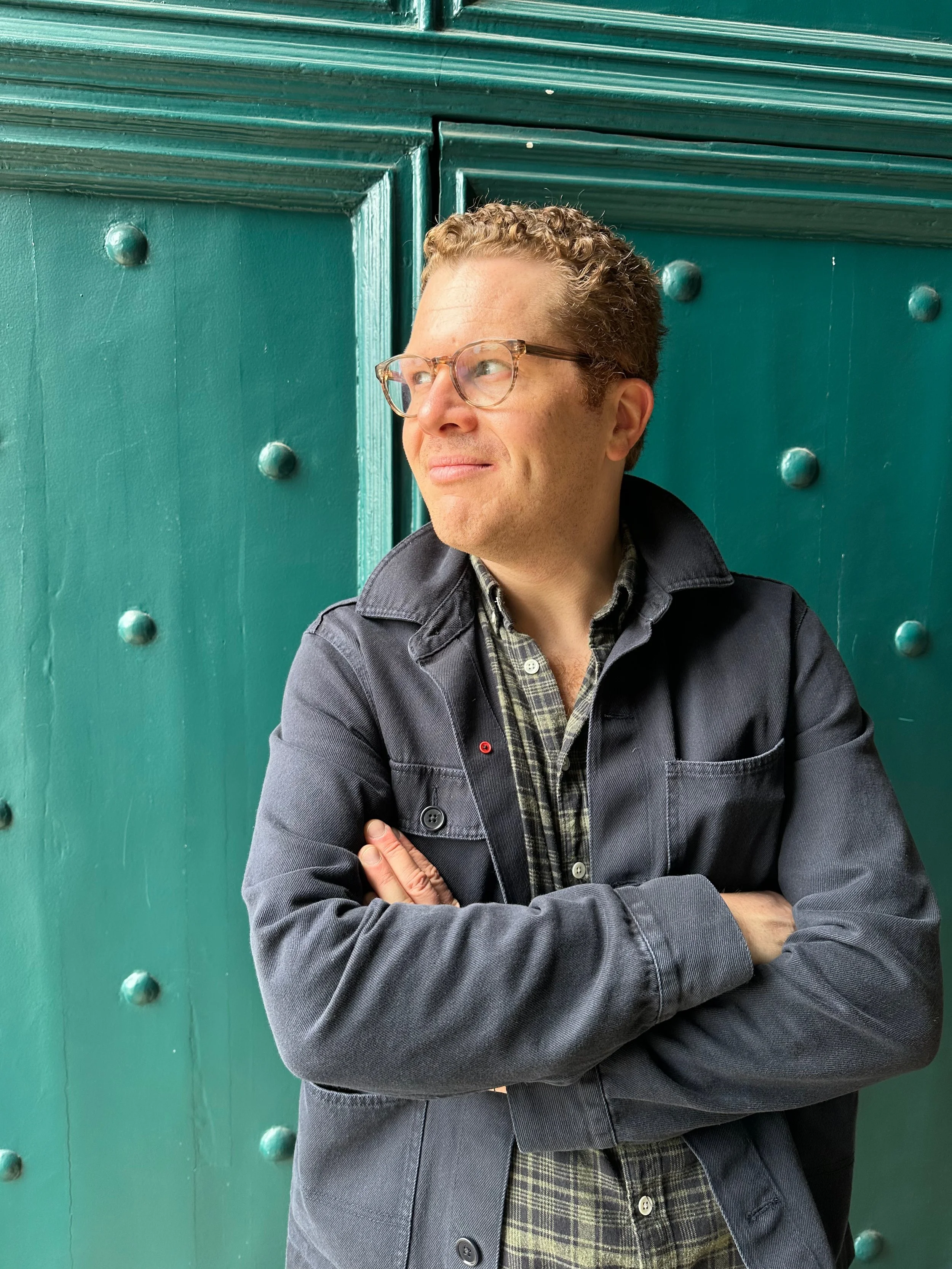 A man with curly hair and glasses crossing his arms, standing in front of a large teal-colored door with decorative rivets.