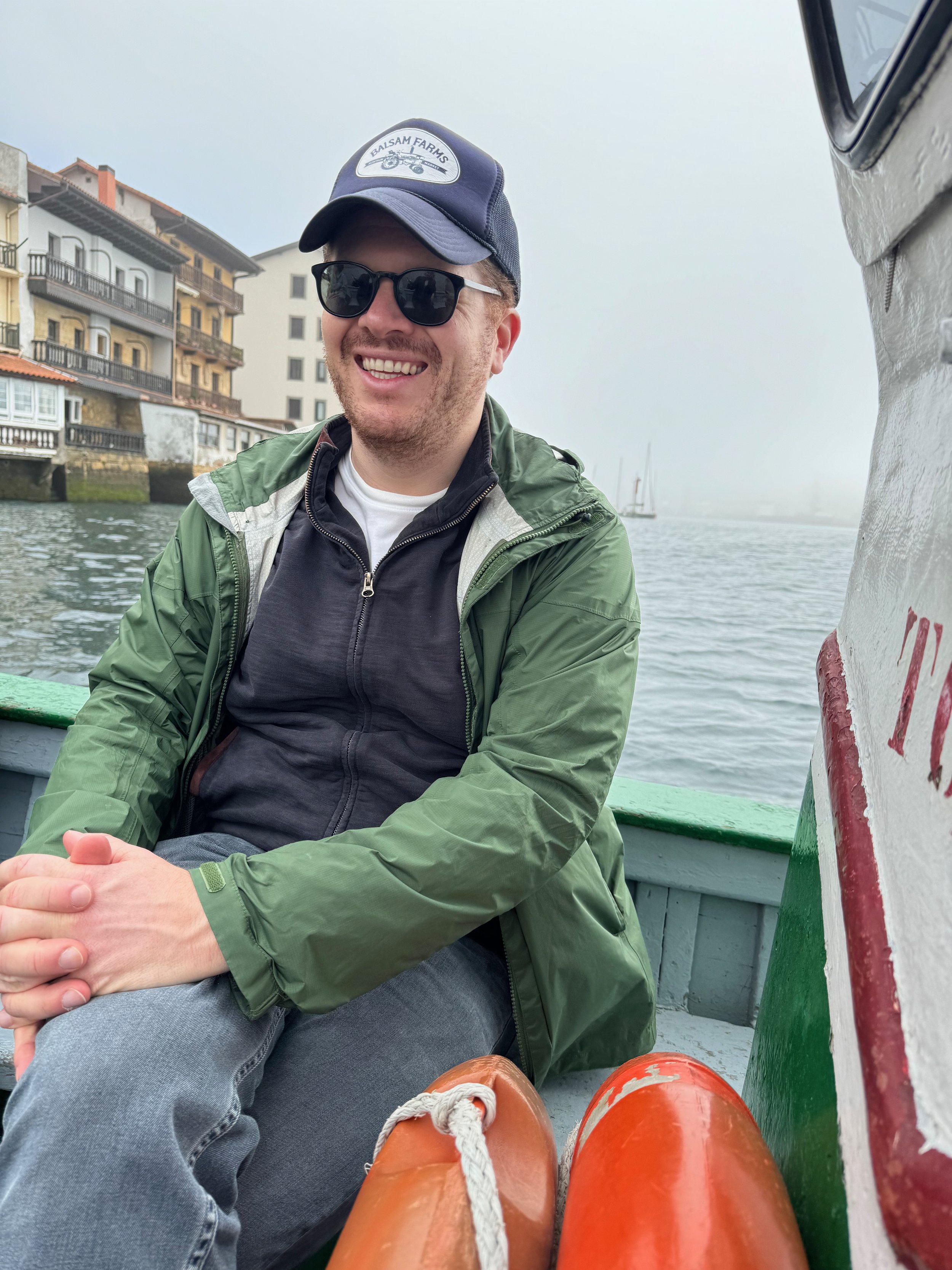 A man smiling, wearing sunglasses, a green jacket, and a navy cap, sitting on a boat with colorful orange and green accents, on a foggy day near waterfront buildings.