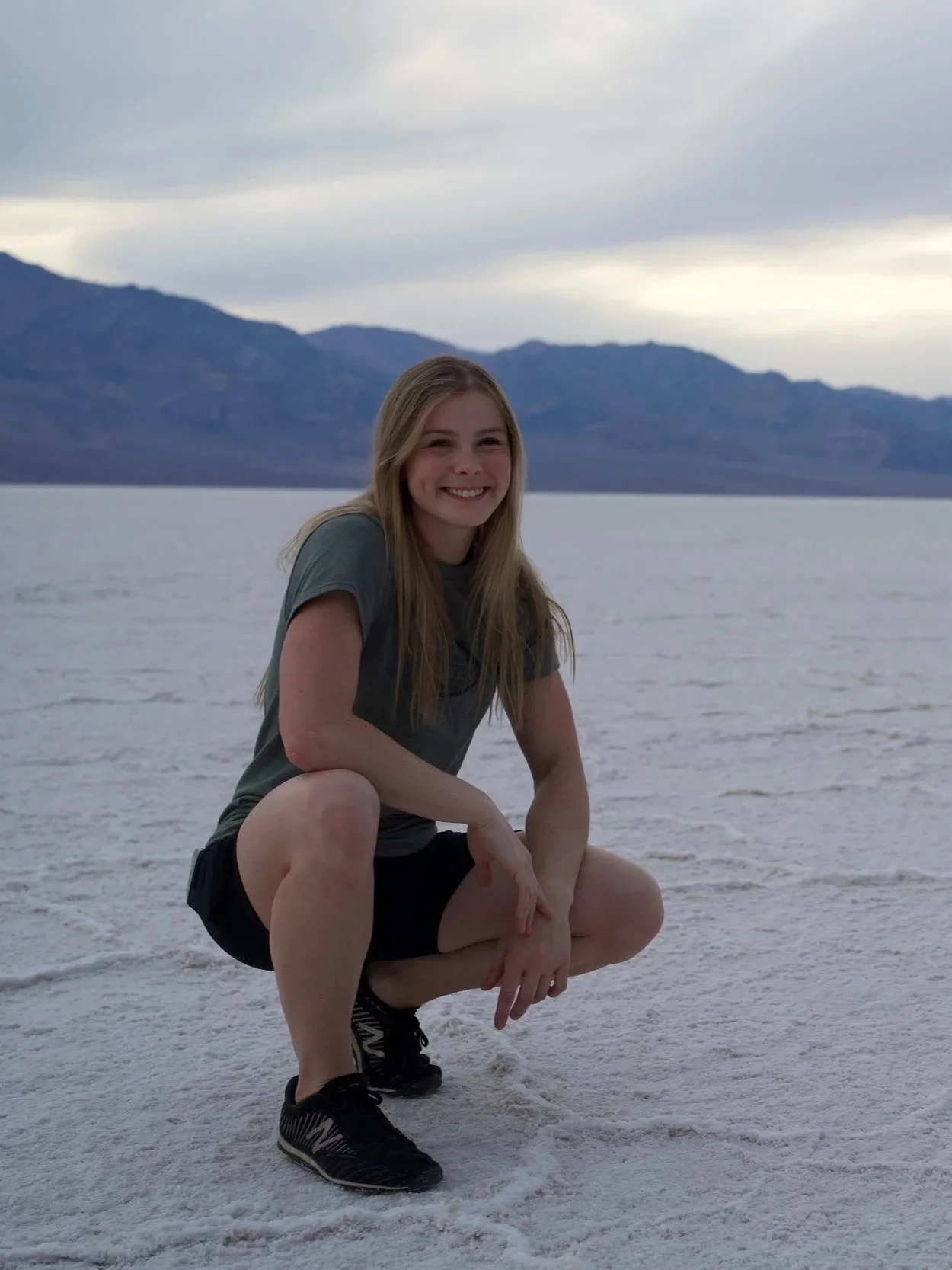 A young woman squatting on a salt flat with mountains and a cloudy sky in the background, smiling at the camera.