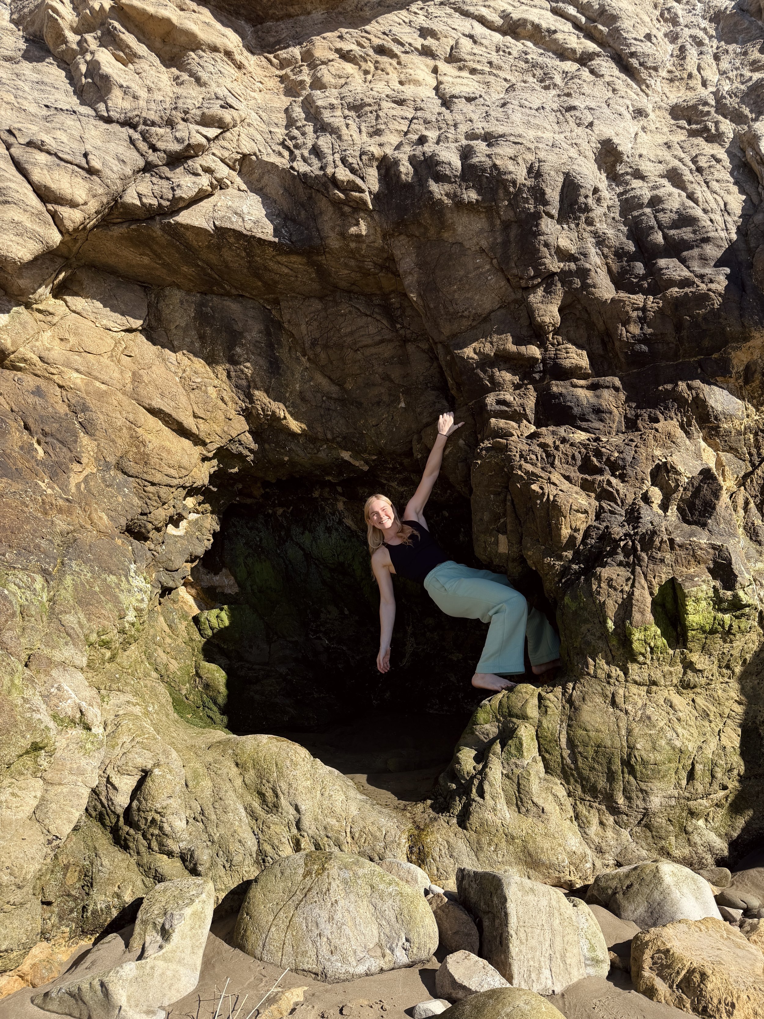 A woman in black top and light pants sitting on rocks in a small cave or rock formation at the beach with sand and large rocks in the foreground.