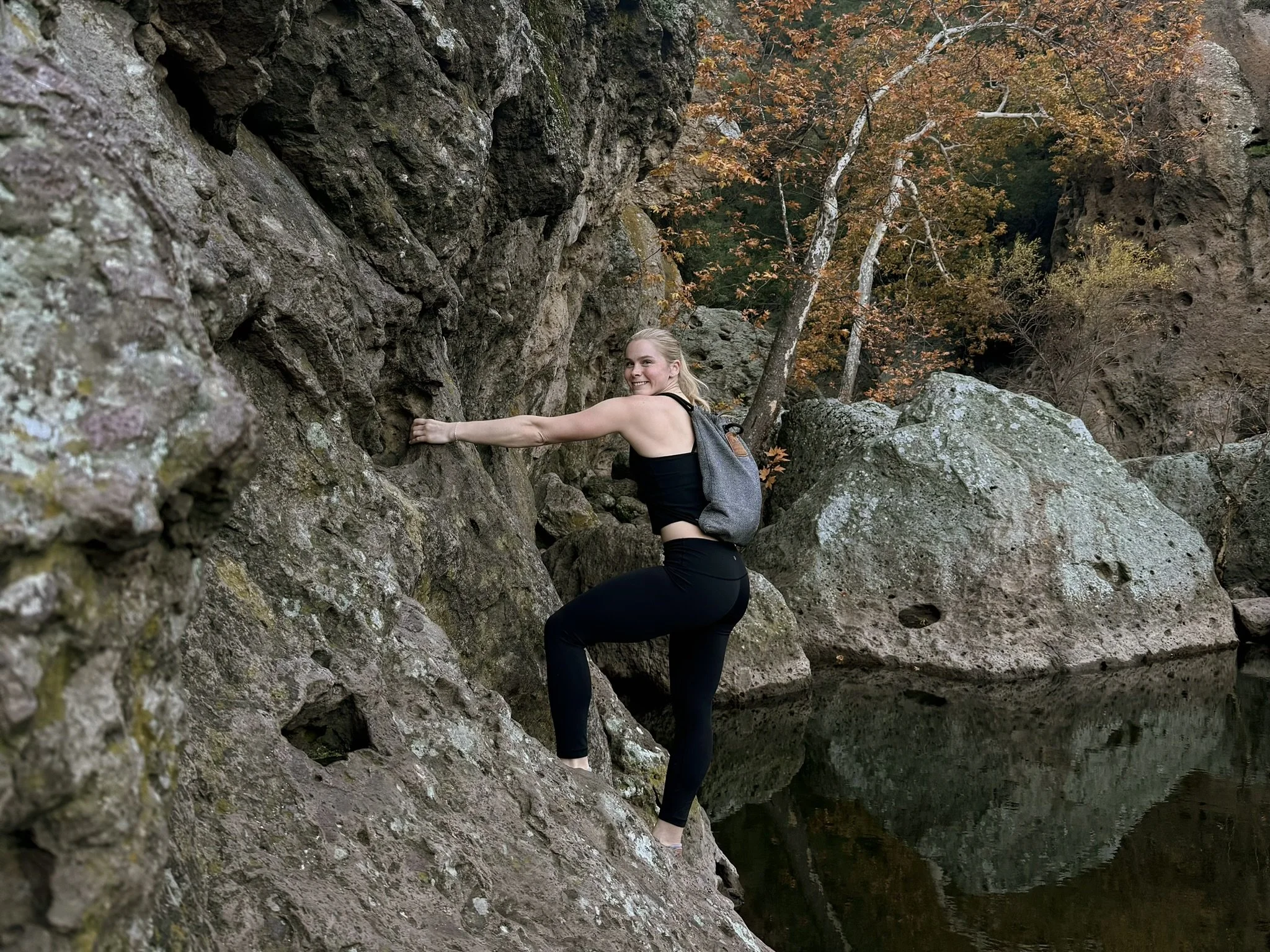 A woman rock climbing on a rugged cliff beside a body of water, with autumn trees in the background.