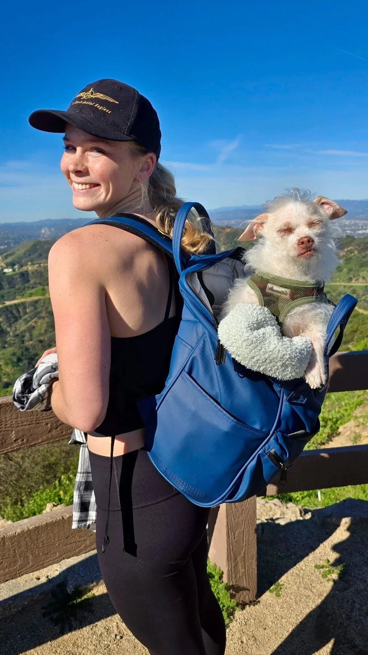 A smiling young woman with a backpack holding a small white dog with closed eyes, wearing a green harness, as they enjoy a scenic outdoor view on a sunny day.