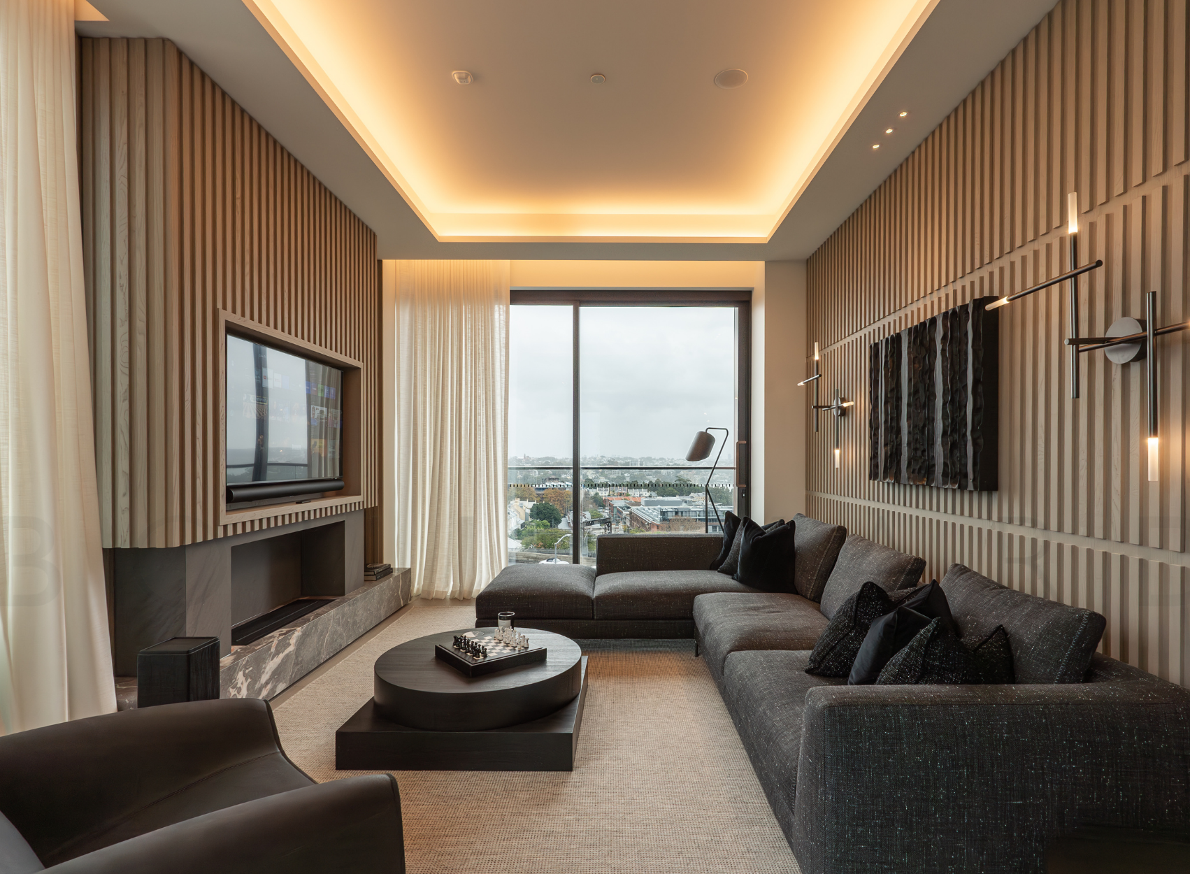 Modern living room with large sectional sofa, coffee table with chess set, wall-mounted television, wood-paneled walls, large window with city view, and contemporary lighting fixtures.