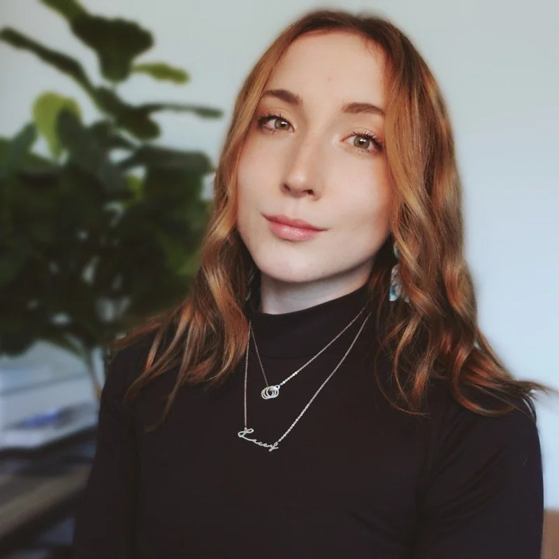 A young woman with light skin and wavy ginger hair wearing a black turtleneck and layered necklaces, standing in front of a blurred green plant and light background.
