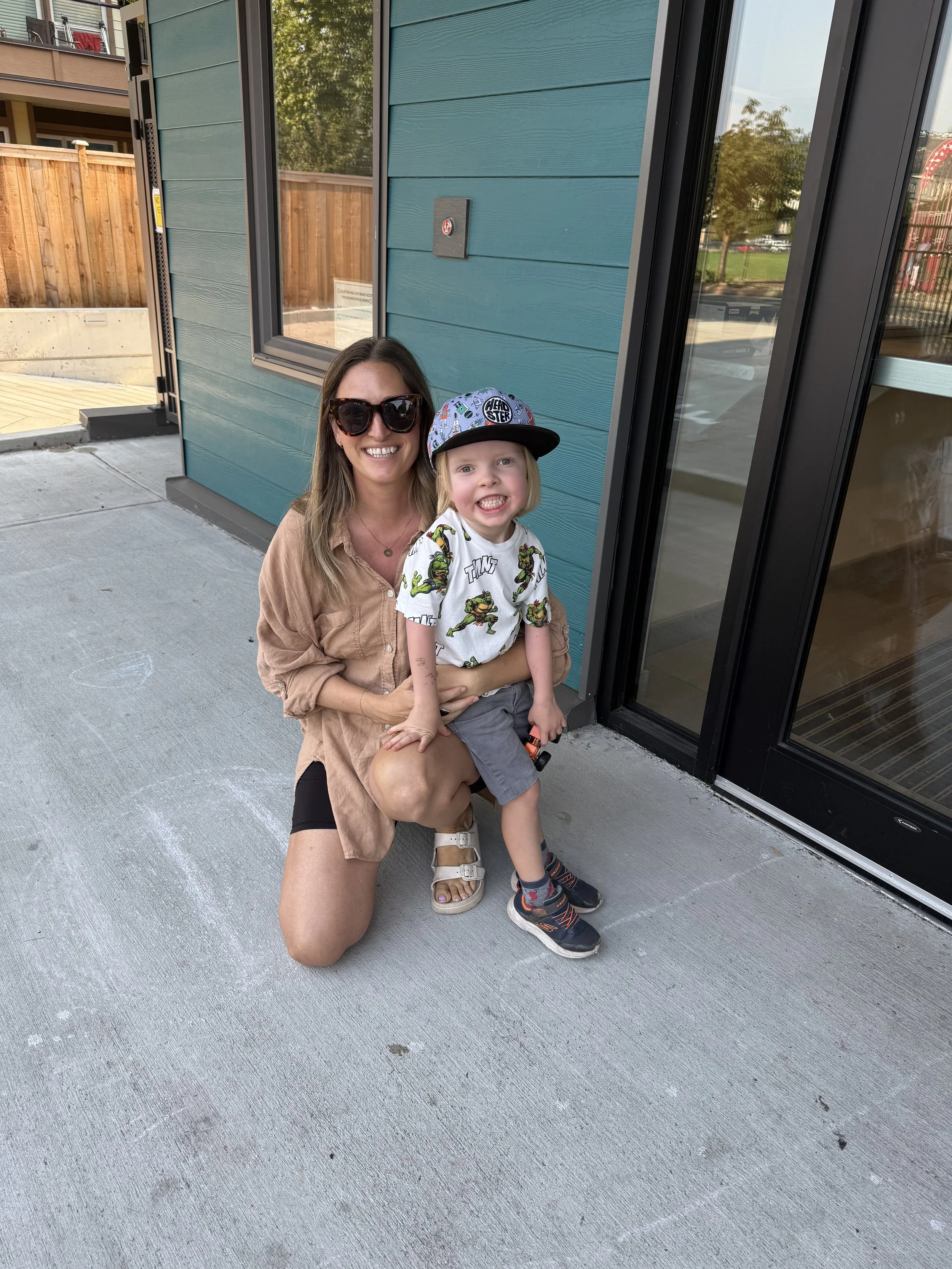 A woman crouching down on a sidewalk next to a young boy who is sitting on her knee. The woman is smiling and wearing sunglasses, while the boy is smiling and showing his teeth. They are near a teal building with a large glass sliding door and a window. There is a wooden fence and trees in the background.