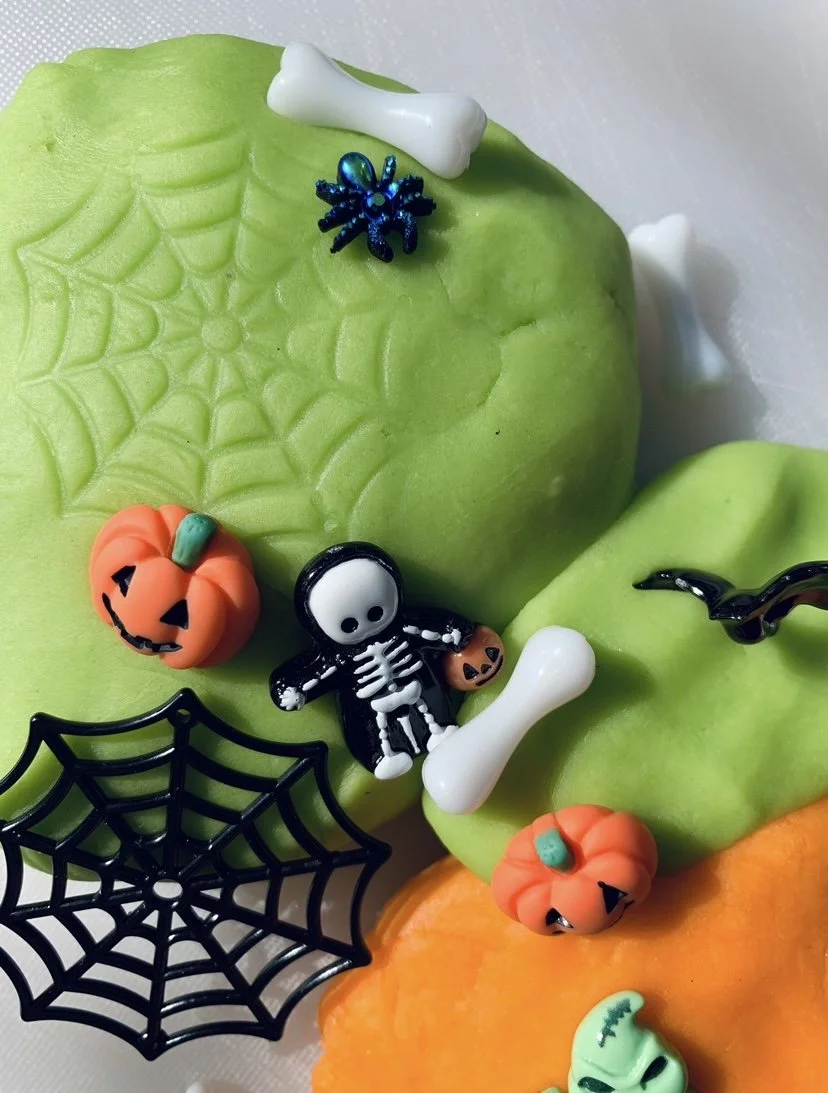 Close-up of a Halloween-themed decorated playdough with green and orange , featuring small Halloween figures including pumpkins, a skeleton, a spider, a black bat, a white bone, a green ghost, and a black spider web.