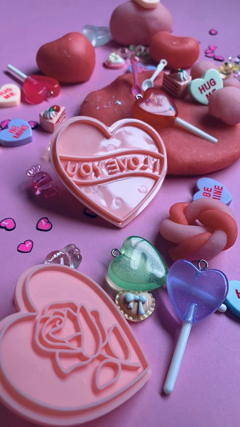 Playdough kit, Close-up of pink hearts, candies, and small toys on a pink background, with a focus on a large pink heart-shaped container with the words "LOVE YOU" embossed on it.