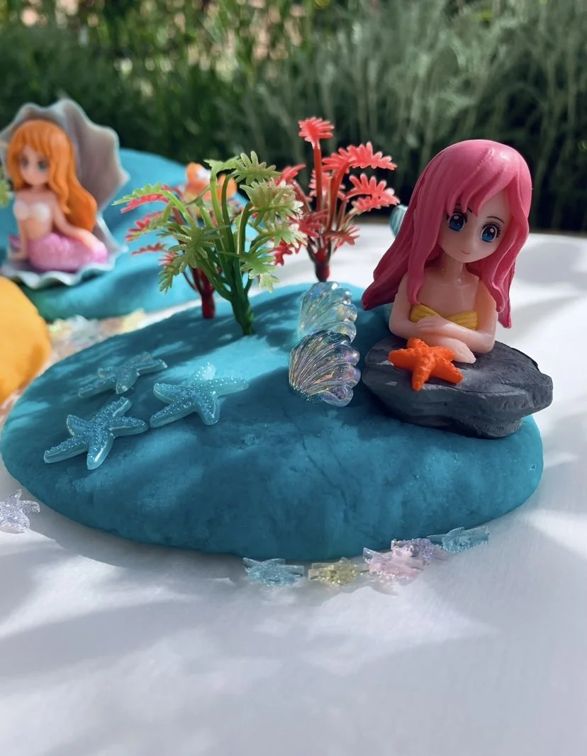 Playdough  scene with a pink-haired mermaid figurine sitting on a black rock in a blue pond, surrounded by plastic plants, starfish, shells, and a small mermaid figurine in the background.