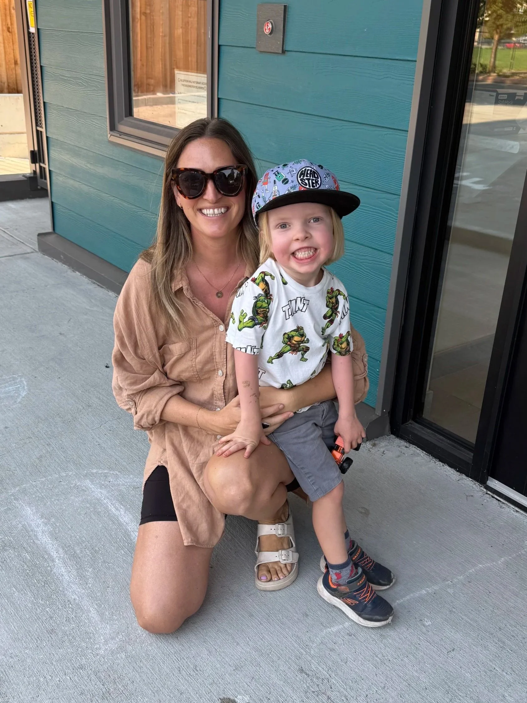 A smiling woman with sunglasses holding a young girl with a baseball cap. They are sitting outside near a blue building with a window and a reflection of a park.