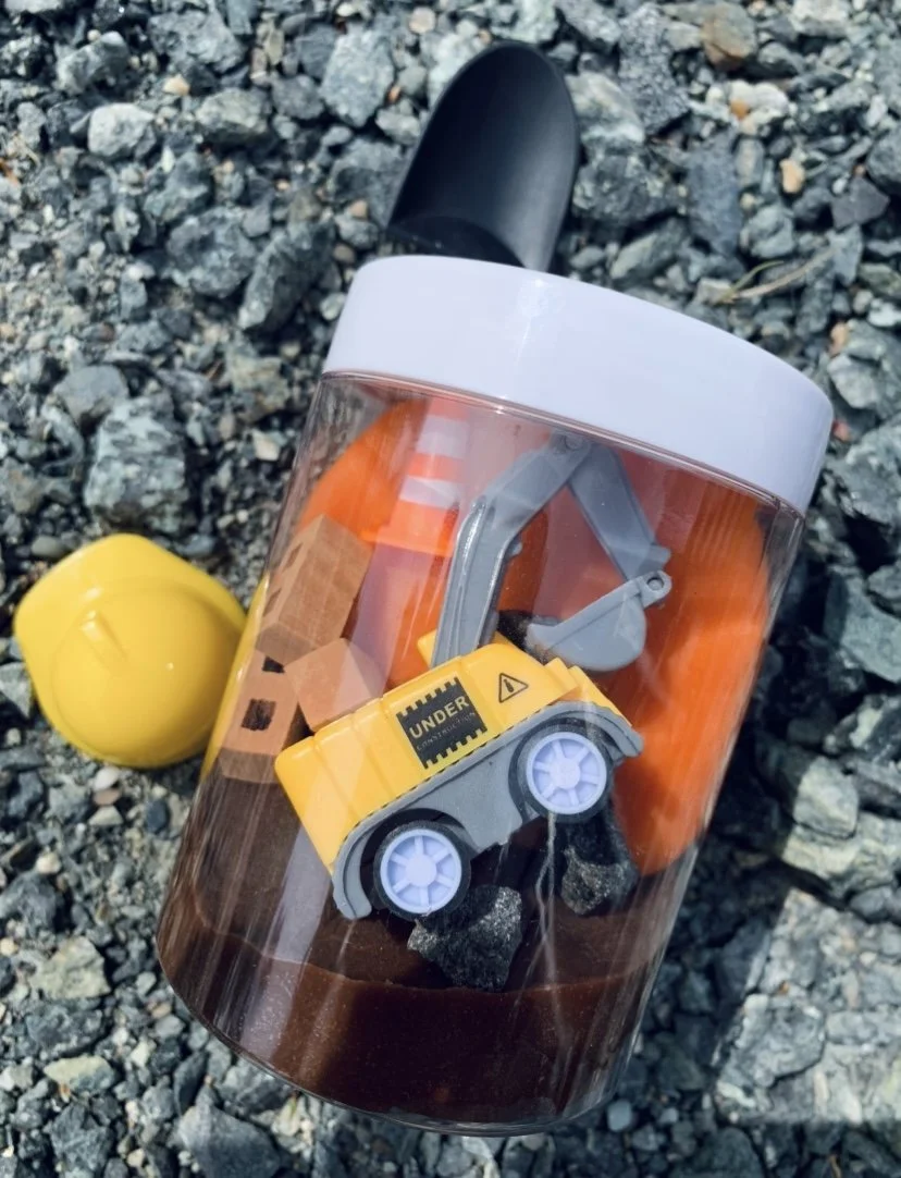 A clear container filled with dirt and toy construction tools and equipment, including a yellow toy excavator, safety cones, and other small plastic tool accessories, peel n stick road a perfect kits play kit.