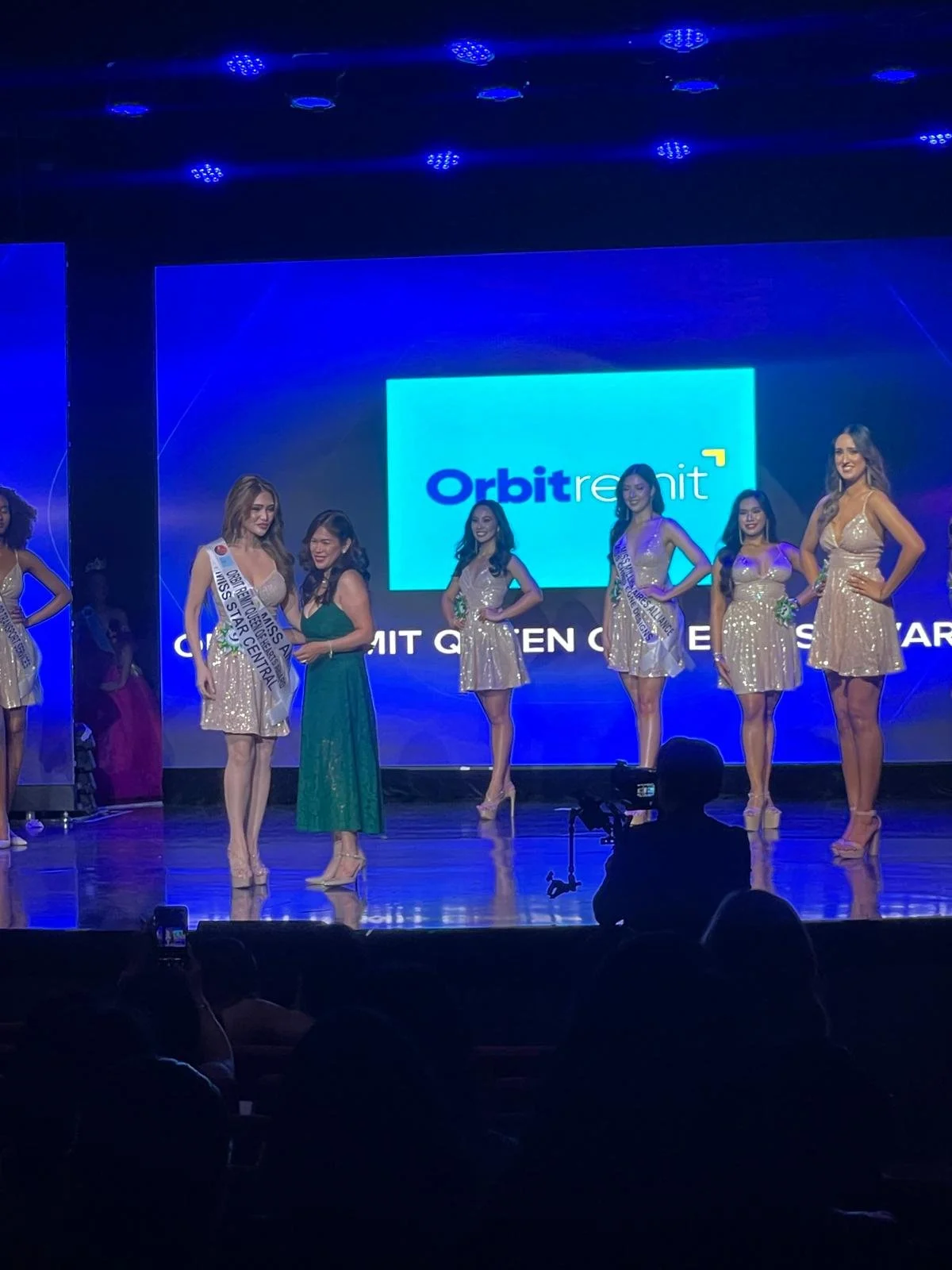 Beauty pageant contestants on stage during an event, with one woman receiving an award, in front of a large screen displaying the Orbit re:mint logo.