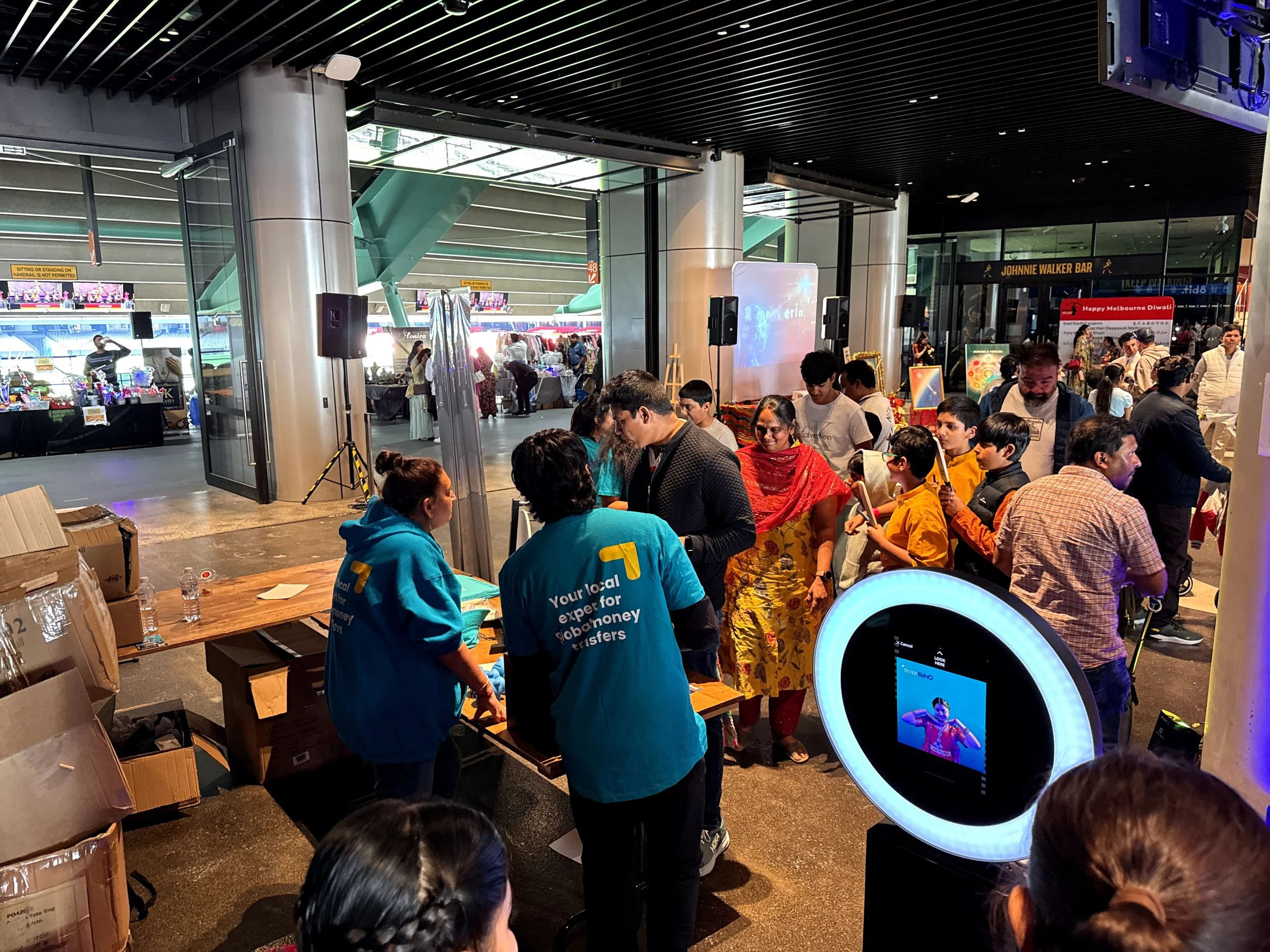 People gathered at an indoor event counter with two people in blue shirts, while a digital camera and a display screen are nearby. In the background, there are other event booths, a stage, and a crowd of attendees.