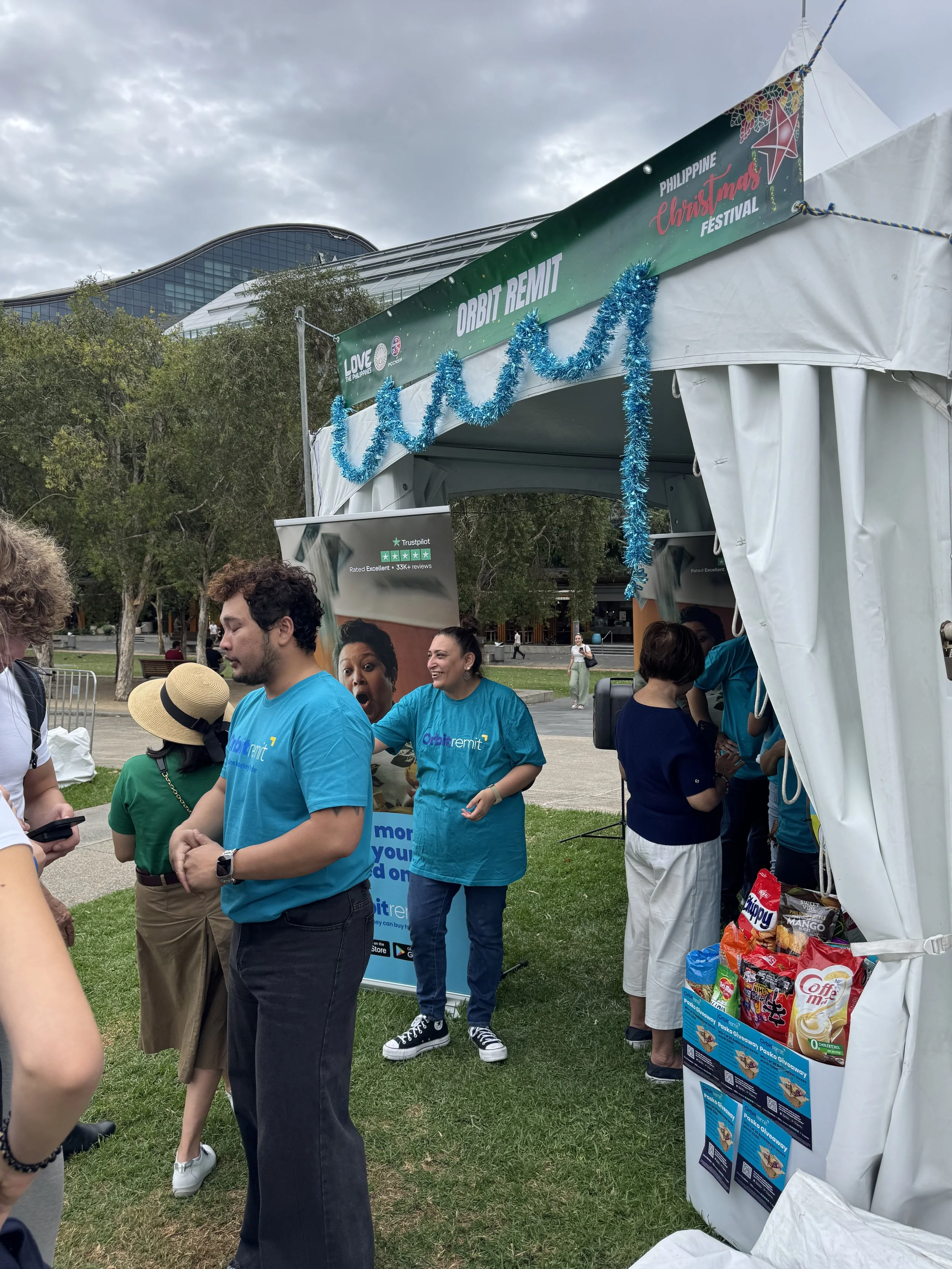People gathered at an outdoor booth during the Philippine Christmas Festival, with a sign reading 'Orbit Remit' and a banner showing the festival name. The booth is decorated with blue tinsel, and snacks are displayed at the side. The background features trees, a modern building, and a cloudy sky.