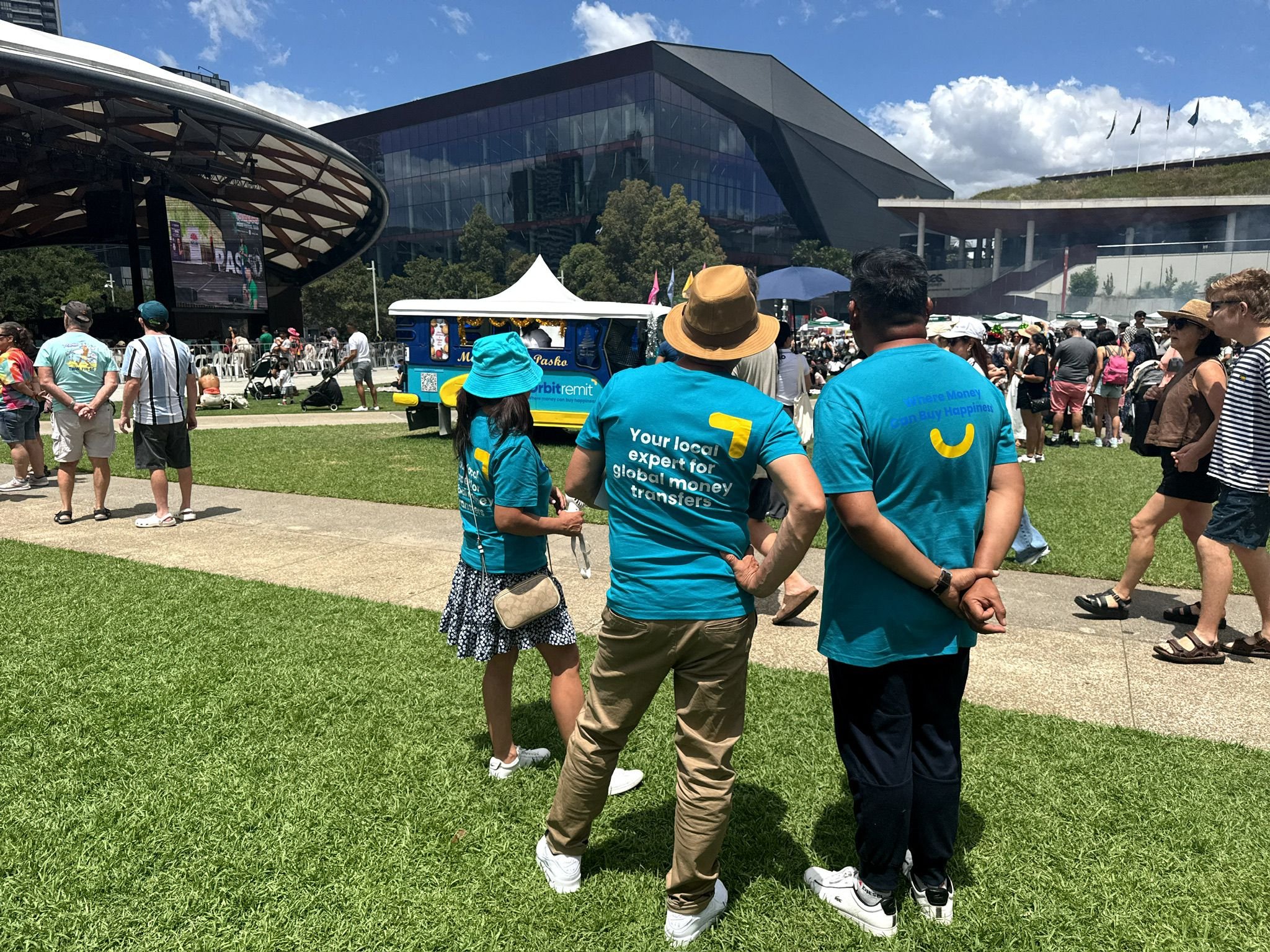 A crowd of people gathered outdoors in front of a modern building with glass and black architecture. Several people are standing on a grassy area, with some wearing blue shirts with logos. There is a blue and yellow tuk-tuk in the foreground, and a large screened stage with people in the background.