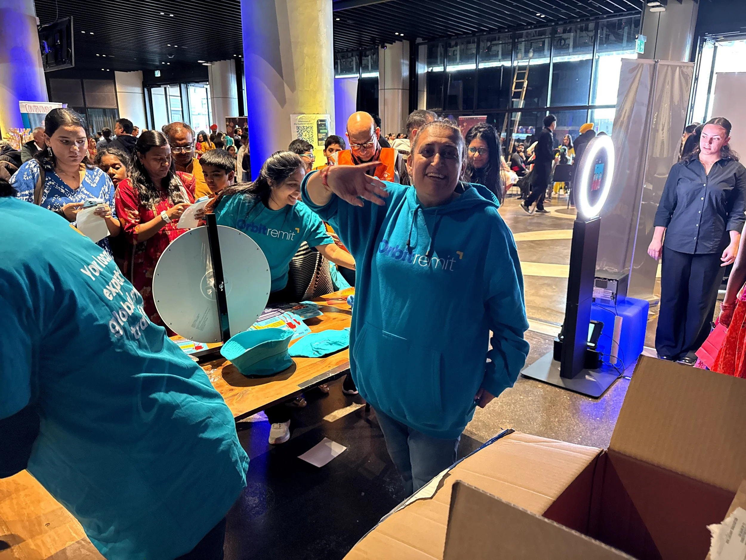 A woman in a blue hoodie with 'Orbit Remit' branding smiling and waving at the camera at a crowded event, with people around her engaging at booths and tables inside a large, modern building.