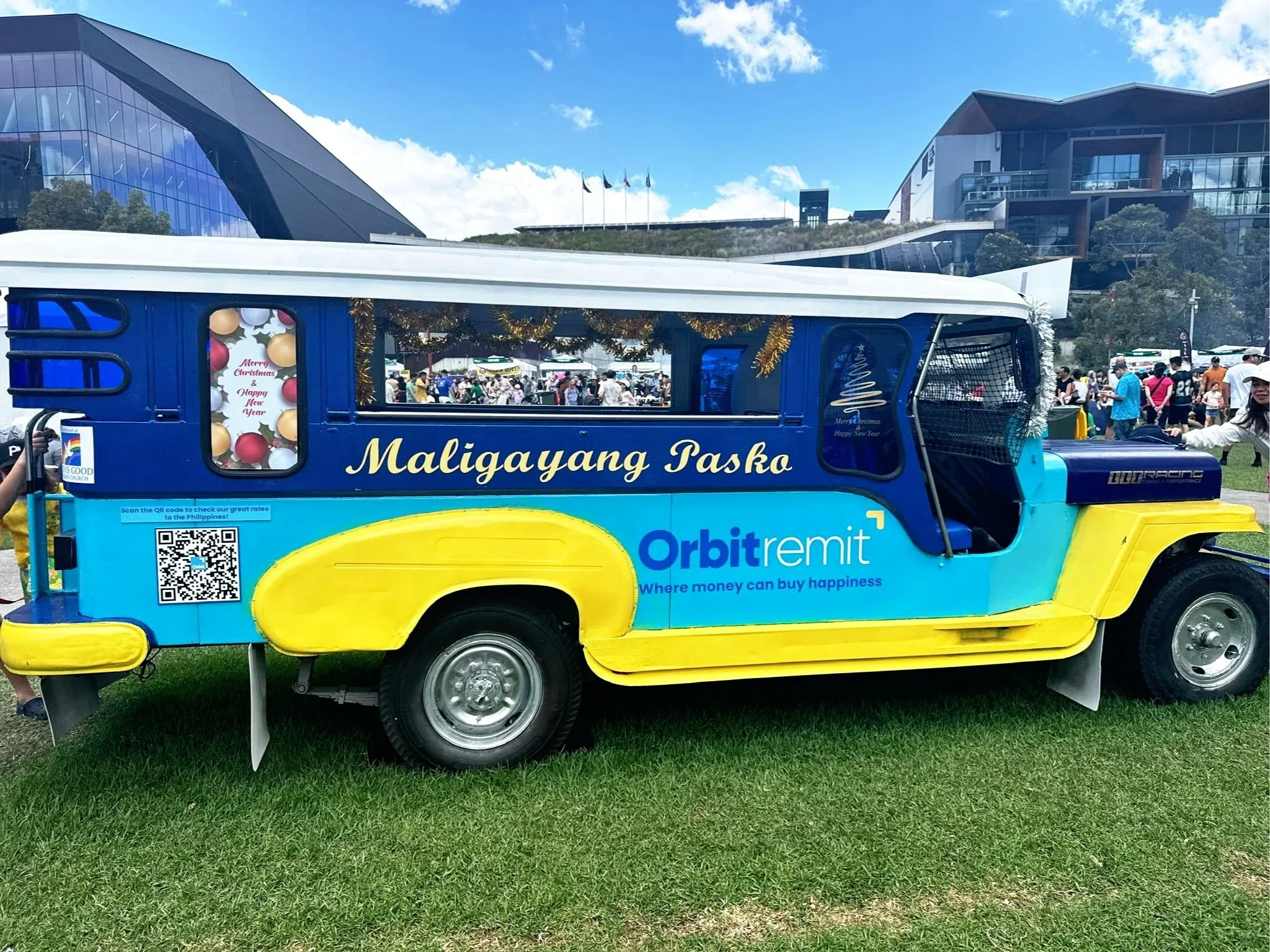 Colorful jeepney decorated with holiday messages and advertisements, parked on grassy area with people and modern buildings in the background.