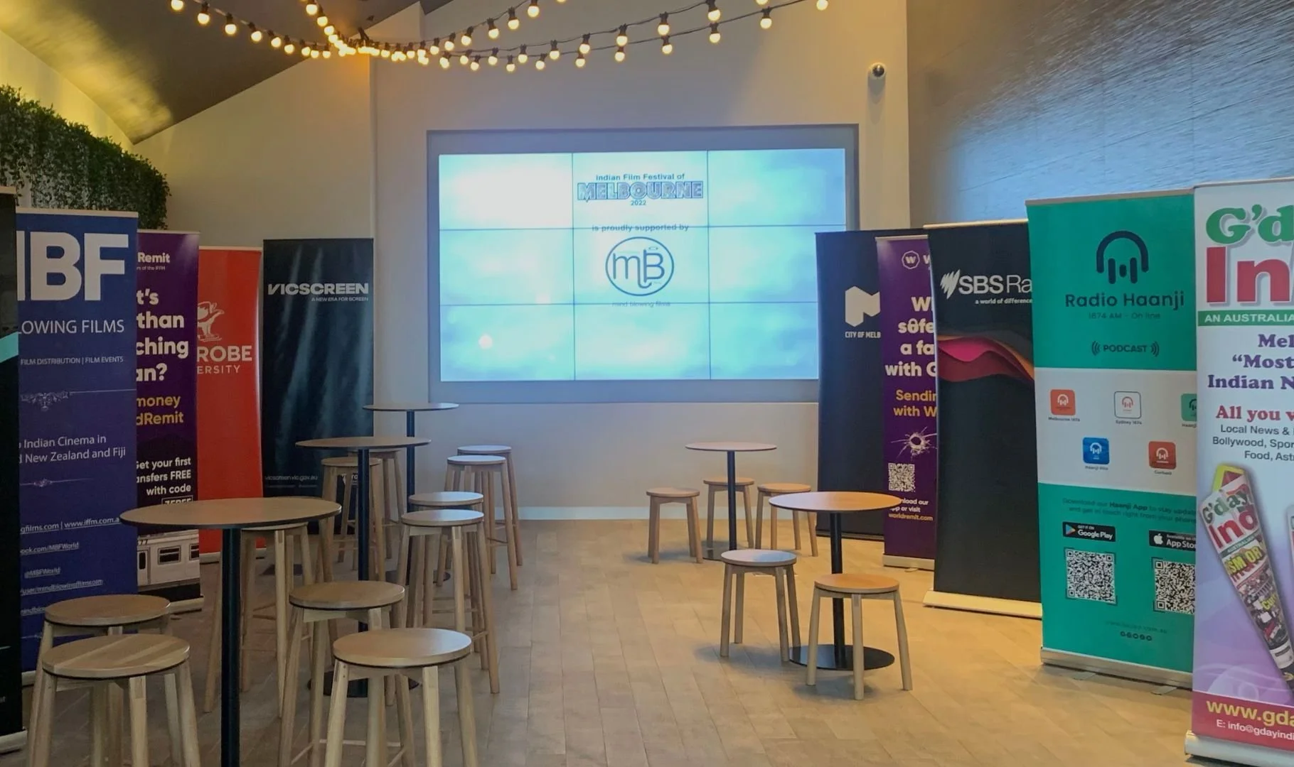 Indoor event space with wooden tables and stools, surrounded by vertical banners for various brands and organizations, and a large digital screen displaying 'Indian Film Festival of Melbourne 2022' with a logo.