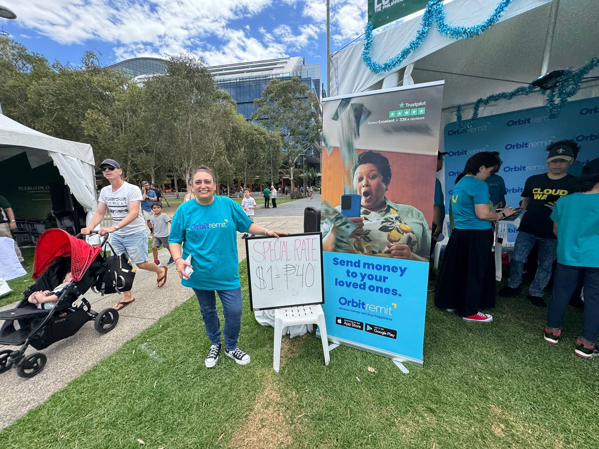 A woman standing next to a sign that reads 'Special Rate $1 = 40' at an outdoor event, with people, tents, and trees in the background. She is wearing a blue OrbitRemit T-shirt and smiling.