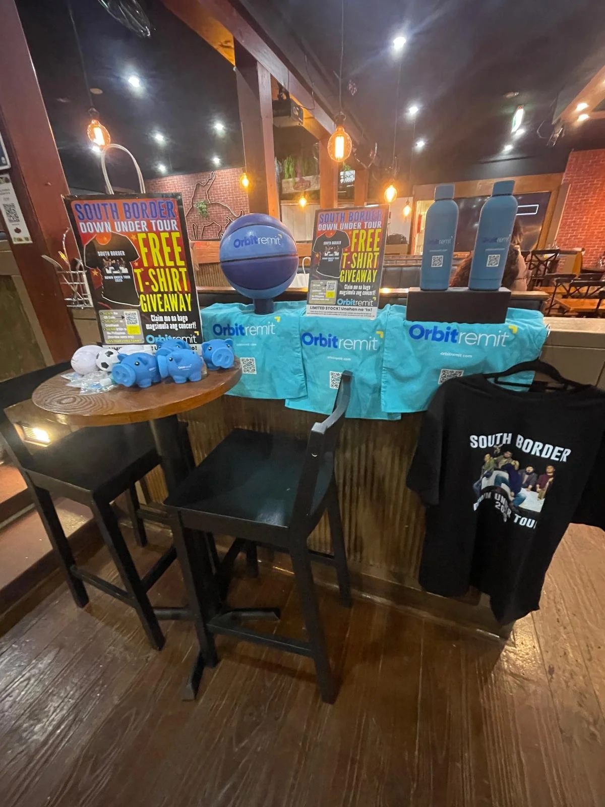 A promotional display table with Orbitremit branding and South Border concert giveaway items including t-shirts, water bottles, and pig-shaped stress balls in a restaurant setting.
