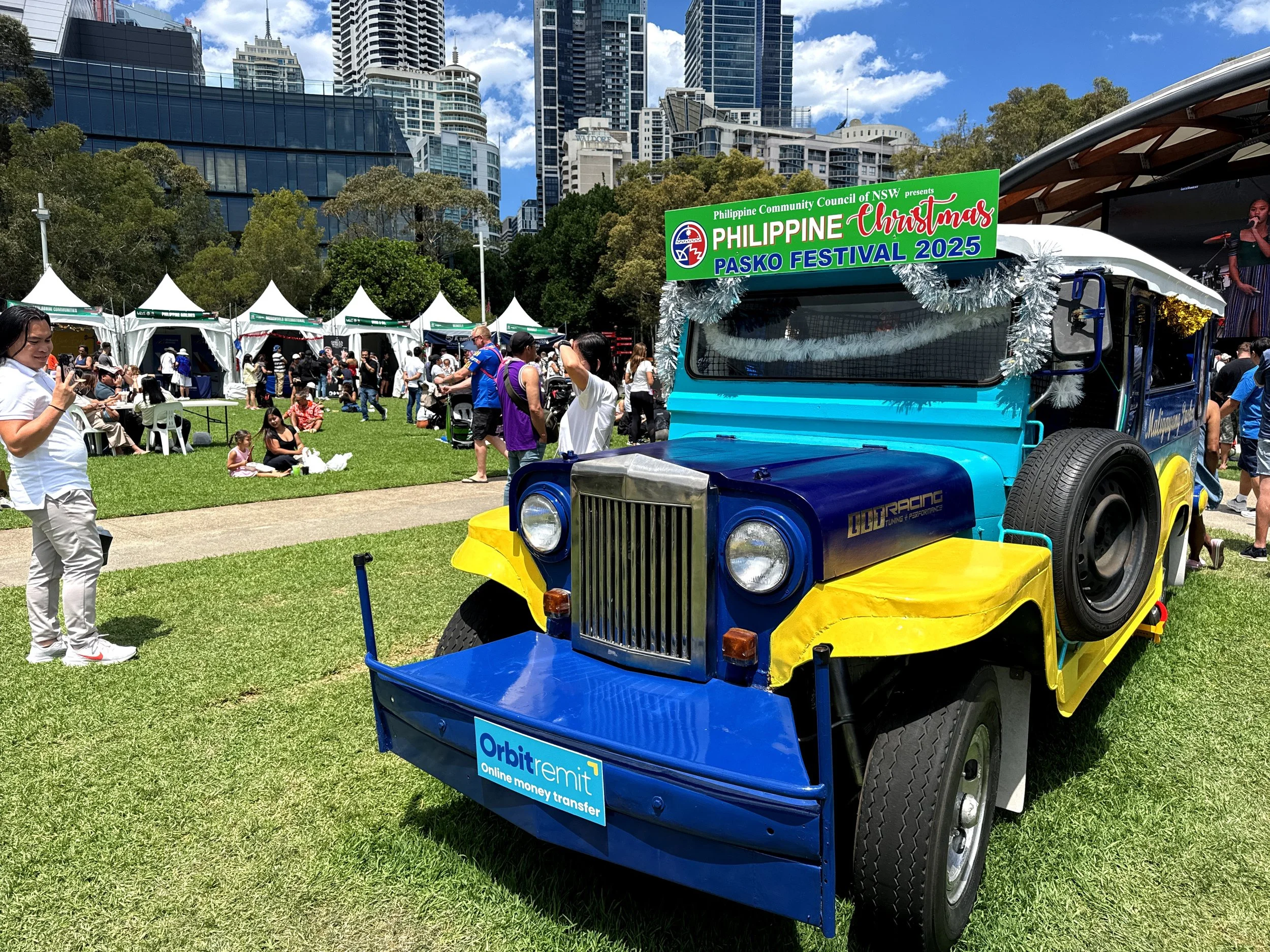 A colorful jeep decorated with tinsel and holiday signs at the Philippine Christmas Pasko Festival 2025, with tents and people gathering in an outdoor park with city buildings in the background.