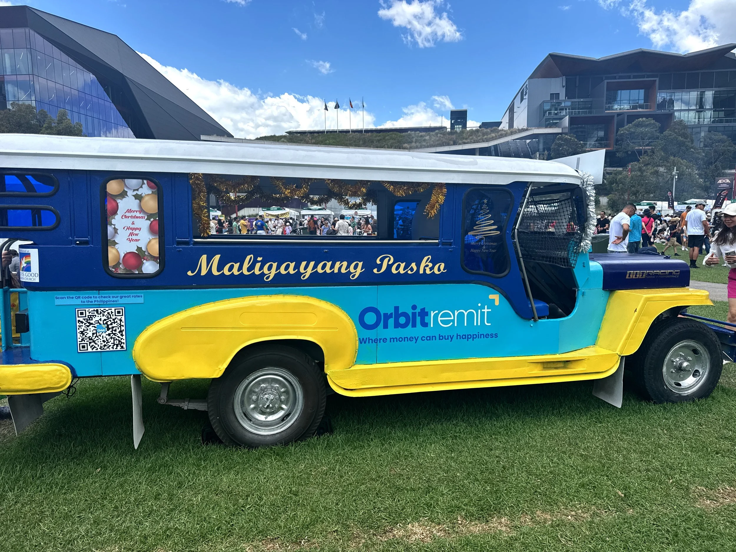 Colorful jeepney decorated for Christmas with gold tinsel and a sign with Philippine text and 'Orbit Remit' logo, parked on grass during a busy outdoor event.