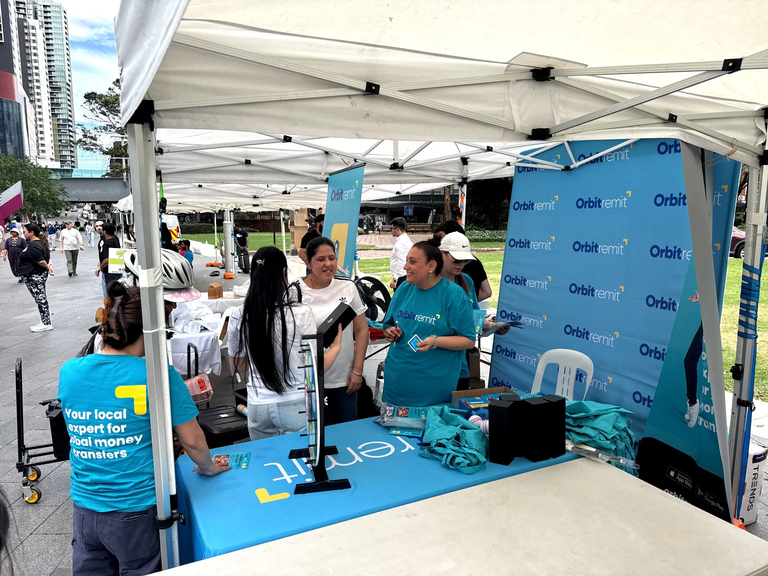 People gathered at an outdoor event booth with blue 'Orbitremit' banners, displaying promotional materials and merchandise, with some wearing branded t-shirts.