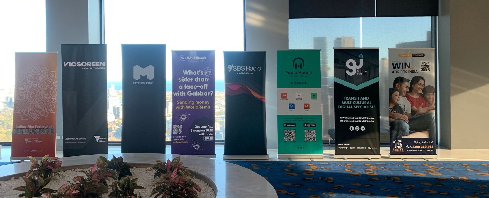 Indoor view of multiple vertical banners advertising various media and travel services, positioned in front of large windows showing a cityscape with tall buildings, with a plant in the foreground and a blue patterned carpet on the floor.