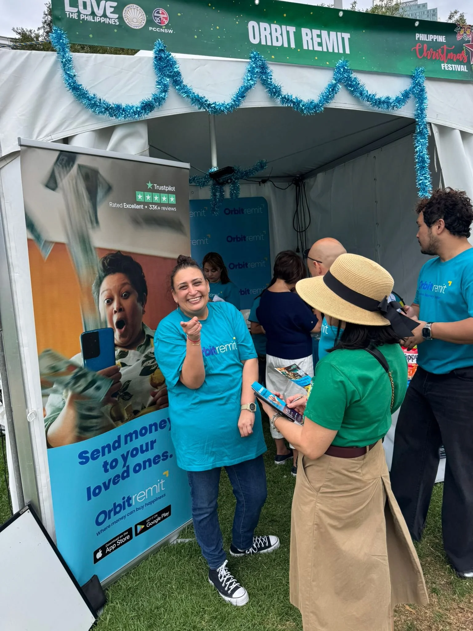 A group of people inside an outdoor booth for Orbit Remit during a Christmas festival. The booth has blue tinsel decorations and an advertisement banner with a smiling woman holding money. One woman in a blue T-shirt is smiling and pointing, while another woman in a green shirt and wide-brimmed hat is looking at her phone. Other people are interacting at the booth.