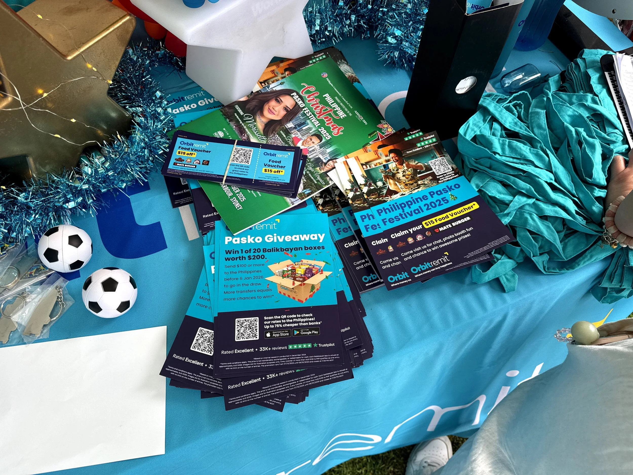 A table displays event flyers, promotional cards, and small soccer ball figures. The flyers promote the Philippines Pasko Festival 2025 and a food voucher giveaway. The table is decorated with blue tinsel, a star-shaped ornament, and some round objects.