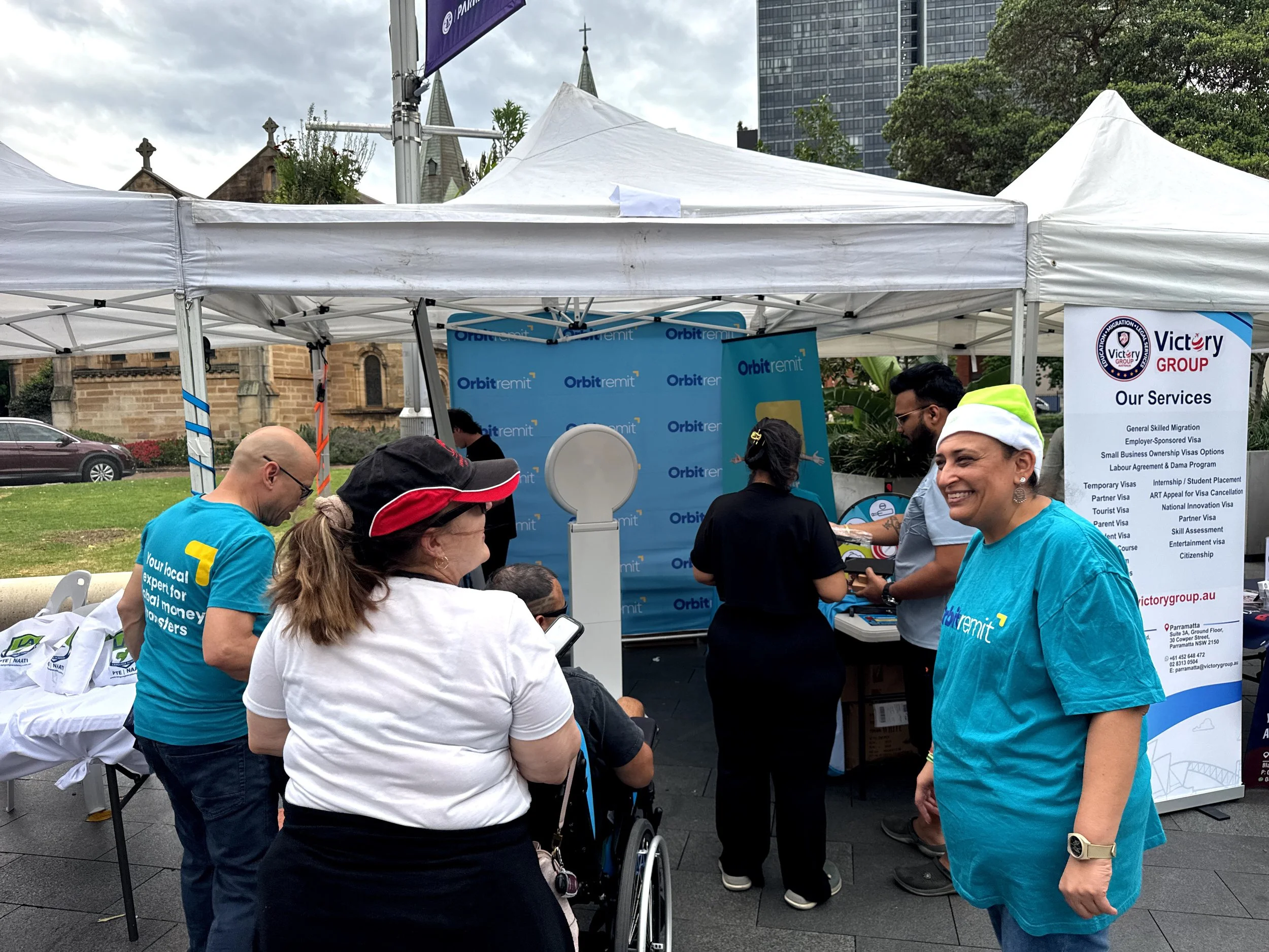 A group of people at an outdoor booth with a blue 'Orbit Remit' banner, showcasing migration and visa services. Some people are standing in line, one woman is smiling, and there are informational posters and flyers on the table.