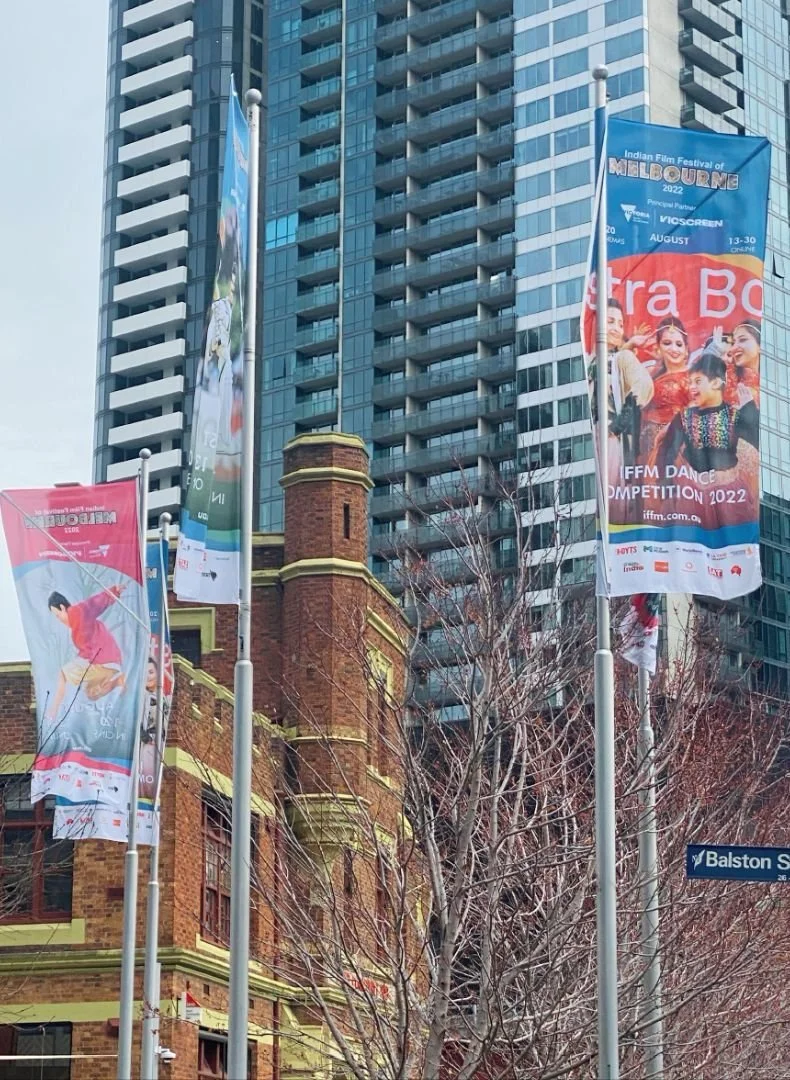 Several colorful flags on poles in front of a historic brick building and a modern glass high-rise skyscraper, with leafless trees and a street sign that says 'Balston S'.