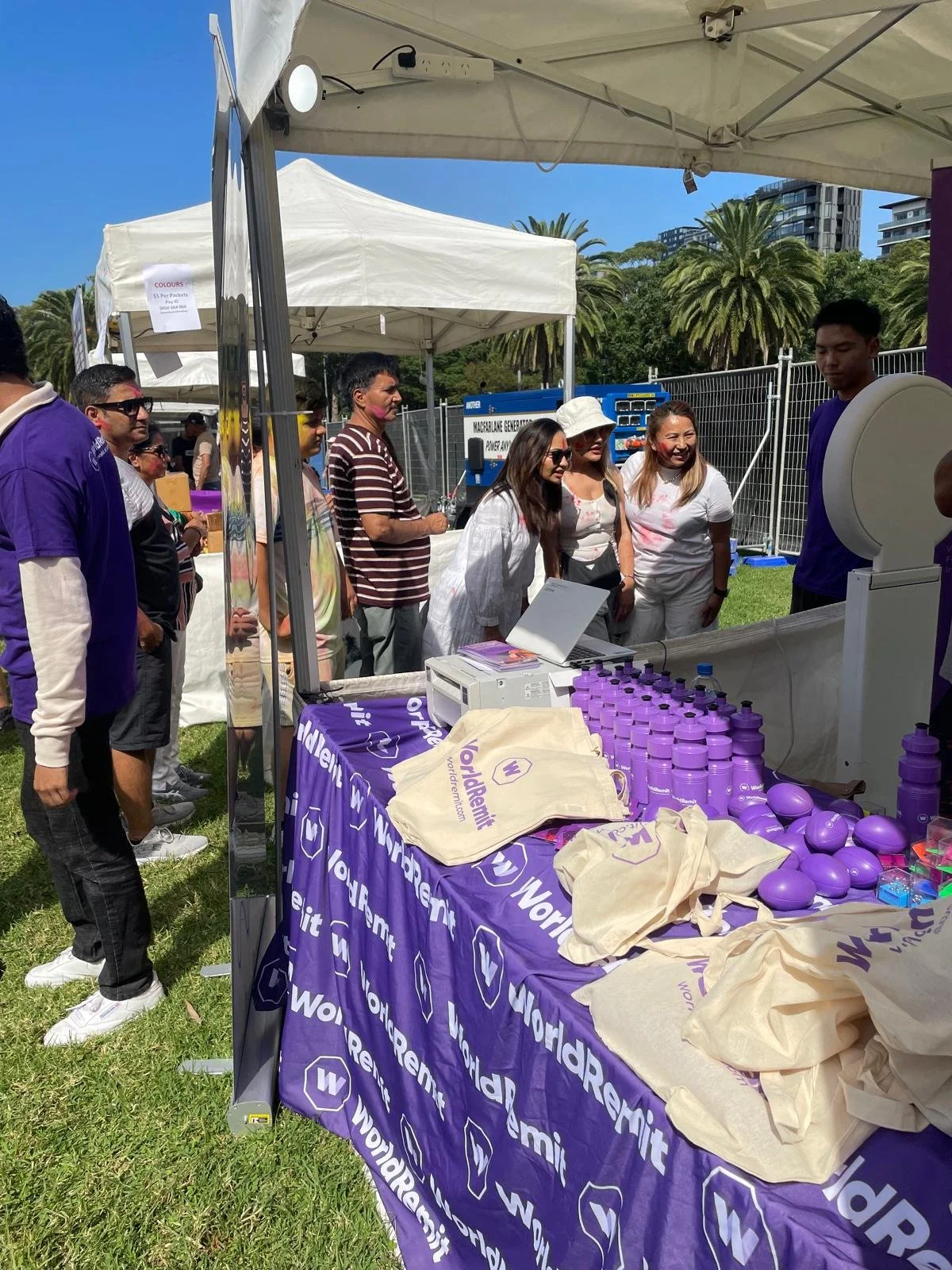 A group of people at an outdoor event gathered around a booth with a purple 'WorldRemit' tablecloth, displaying purple water bottles and promotional items, under white tents with palm trees and high-rise buildings in the background.