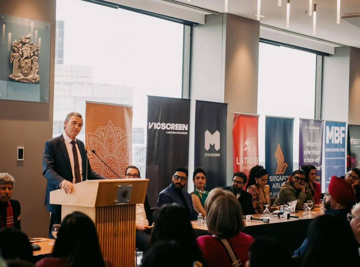 A man in a suit speaking at a podium during a panel discussion at a conference, with several seated panelists and banners from various organizations behind him in a modern room with large windows.