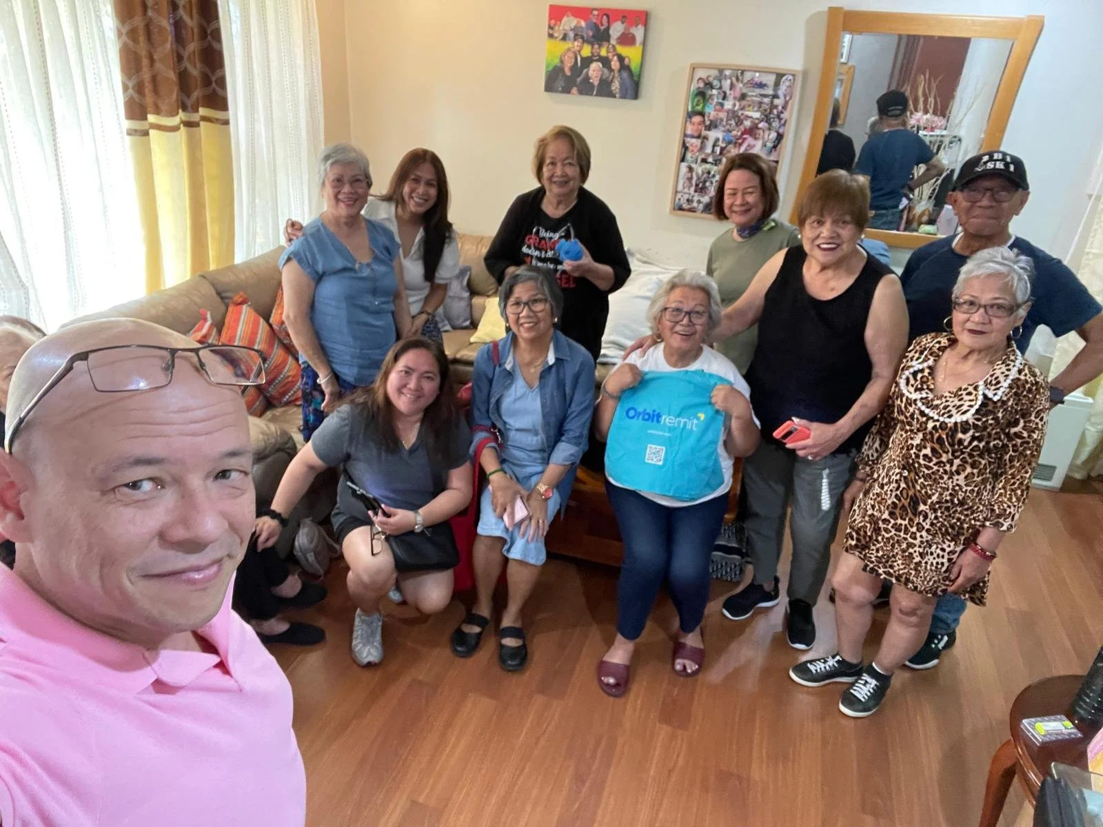 A group of fifteen people, mostly women, gathered in a living room, smiling and posing for a photo. The person taking the selfie is a bald man wearing glasses on his head and a pink shirt. The room has hardwood floors, a beige sofa with colorful cushions, and wall decorations including photos and artwork.