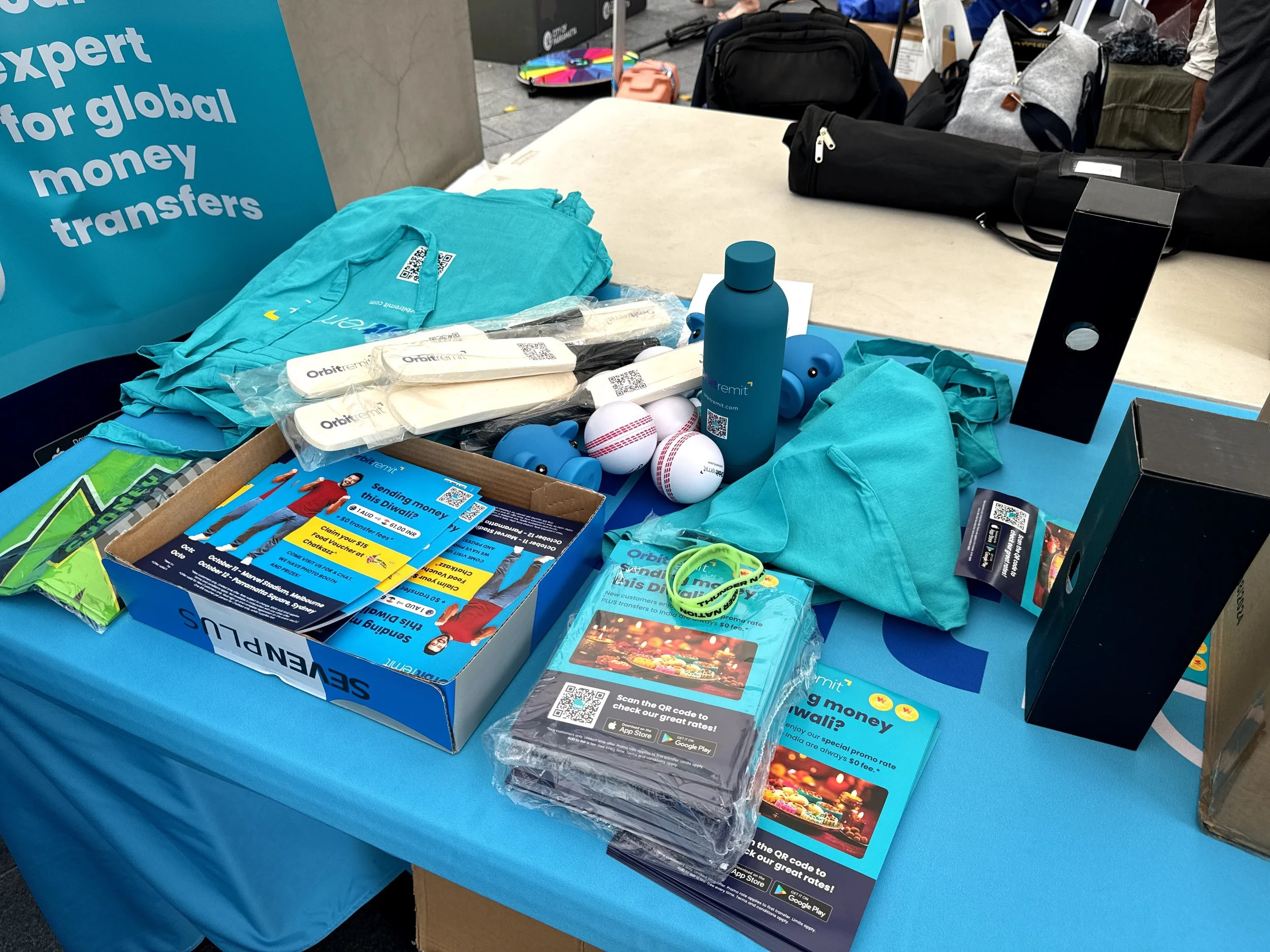 Table with promotional materials, including flyers, wristbands, and small toys for a money transfer service display at an event. Items include branded water bottles, balls, and informational leaflets.