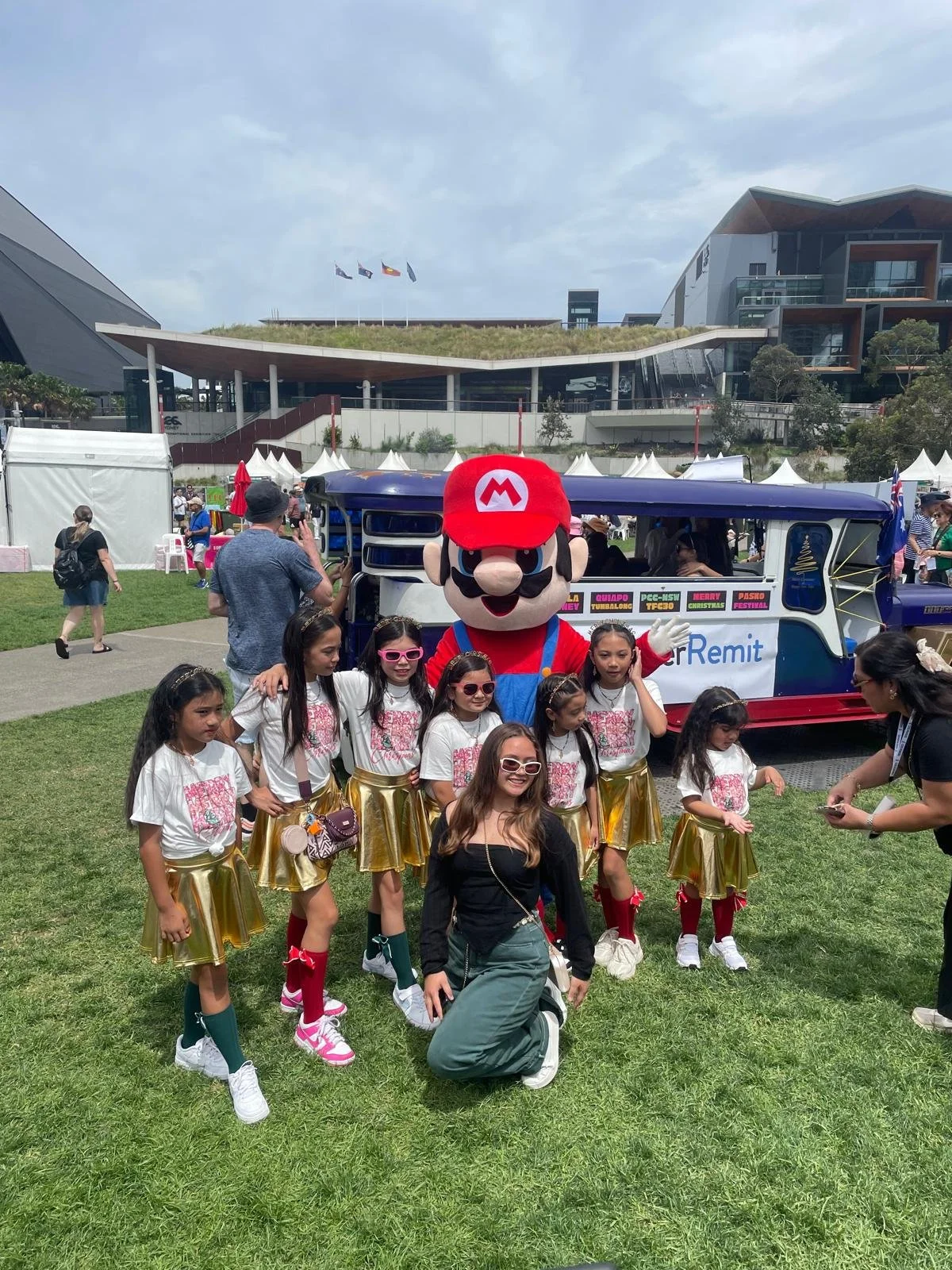 Group of young girls in matching white T-shirts and gold skirts posing with person in Mario costume at outdoor event.