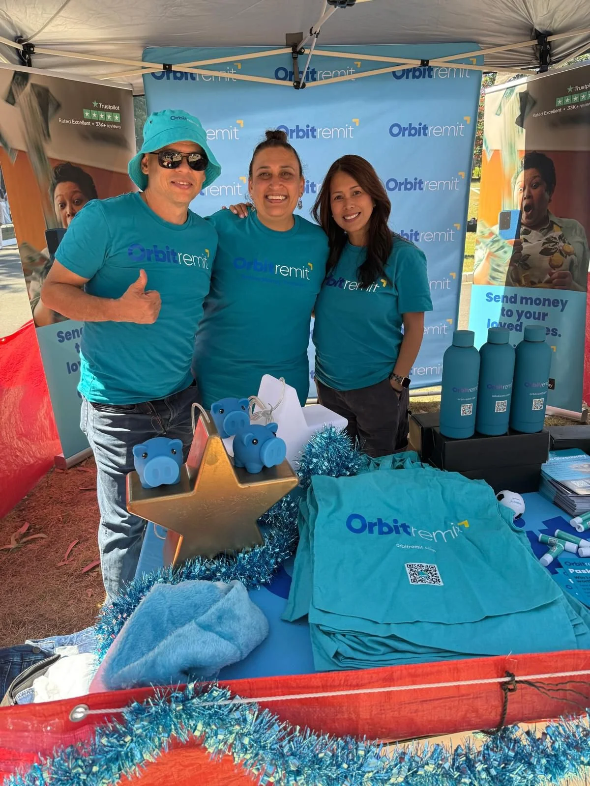 Three people wearing blue Orbit Remit shirts at an outdoor booth, smiling and posing for the photo, with promotional materials and piggy banks on the table.