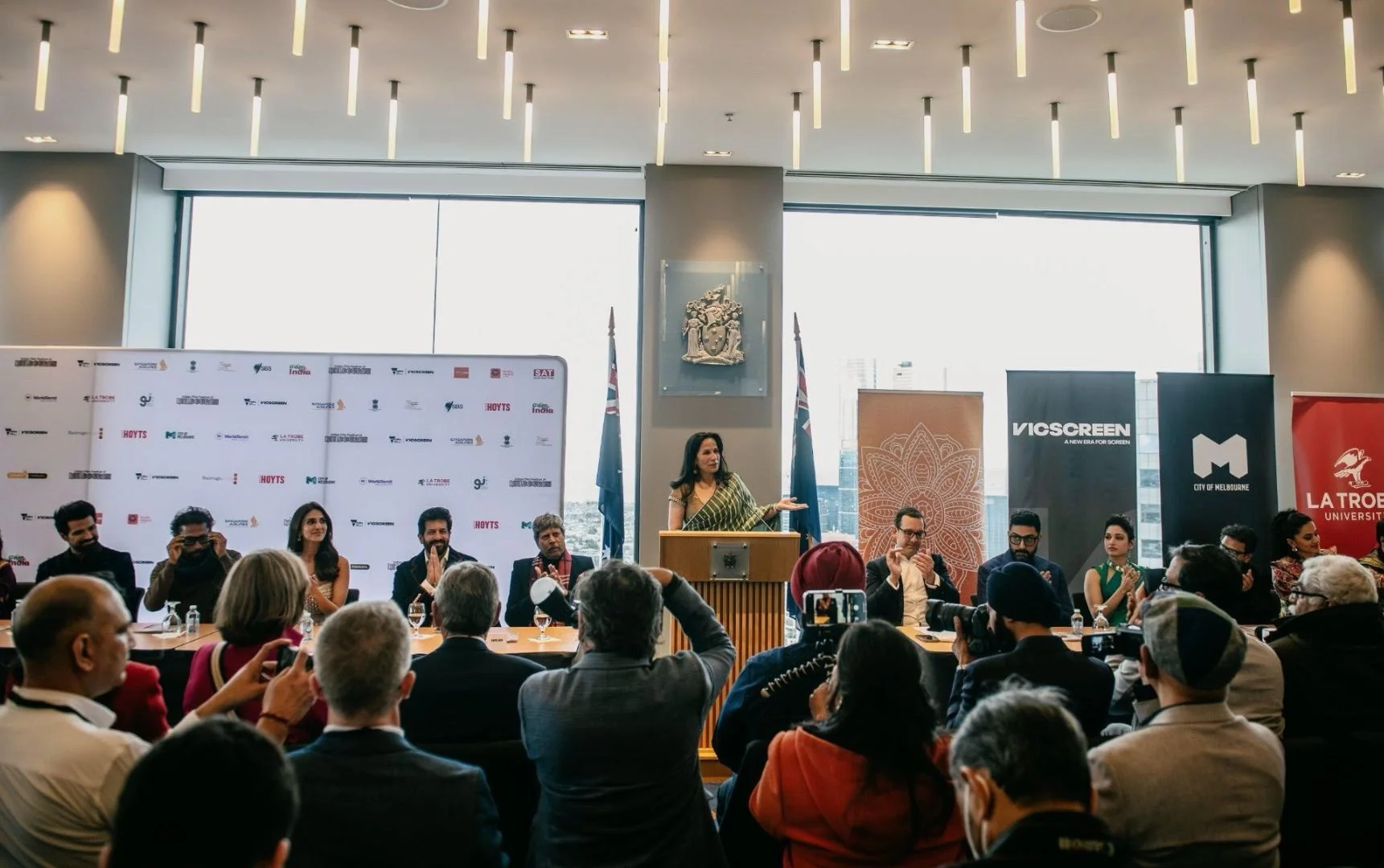 A woman in a green sari speaking at a podium during a conference, with a panel of men and women seated behind her, and audience members listening and taking photos, in a modern conference room with large windows and banners.