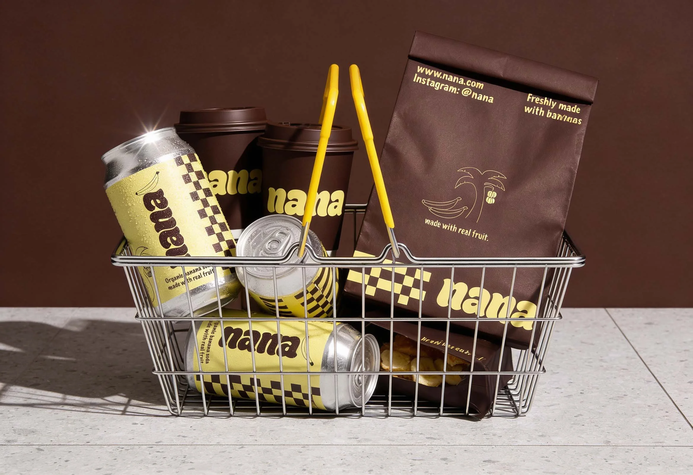 A small shopping cart filled with canned beverages and brown paper bags branded with Nana, placed on a floor against a brown wall.