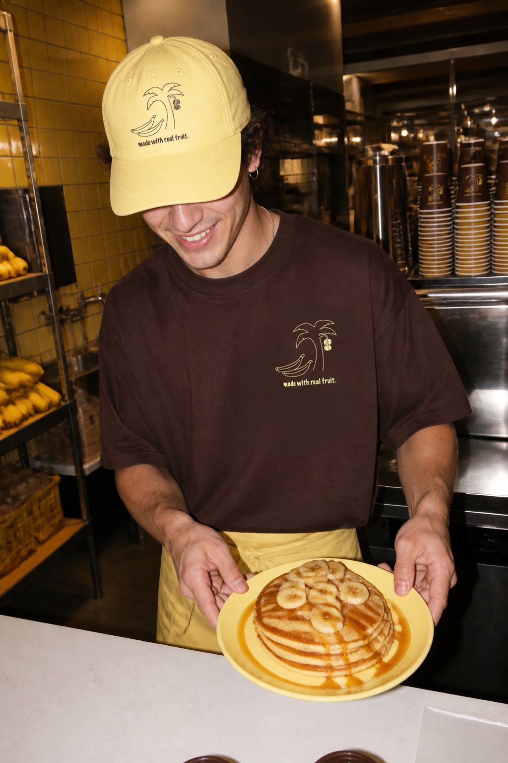 A man wearing a yellow cap and brown shirt with a palm tree and banana logo, smiling and holding a plate of pancakes topped with banana slices and drizzled with syrup, in a kitchen or restaurant setting.