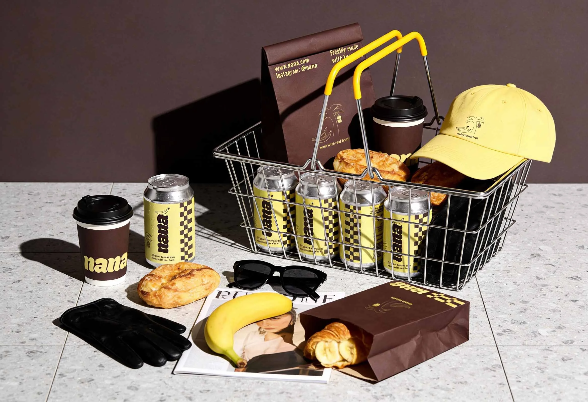 A shopping cart on a table filled with cans of Nana organic banana soda, two paper coffee cups, a yellow baseball cap, sunglasses, magazines, pastries, a banana, and brown paper bags, with a pair of black gloves and a magazine with a photo of a woman on it nearby.