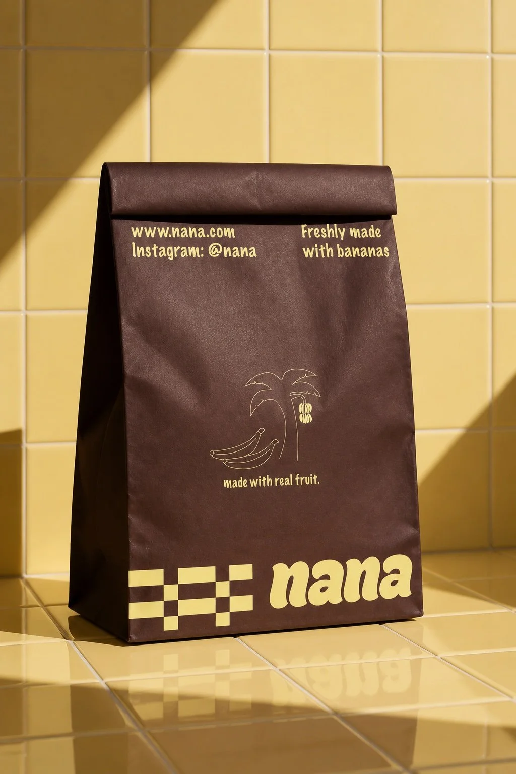 A brown paper bag with yellow text and graphics, sitting on a yellow tiled surface. The bag features a website, Instagram info, a palm tree with bananas graphic, and mentions that it is made with real fruit, created by Nana.