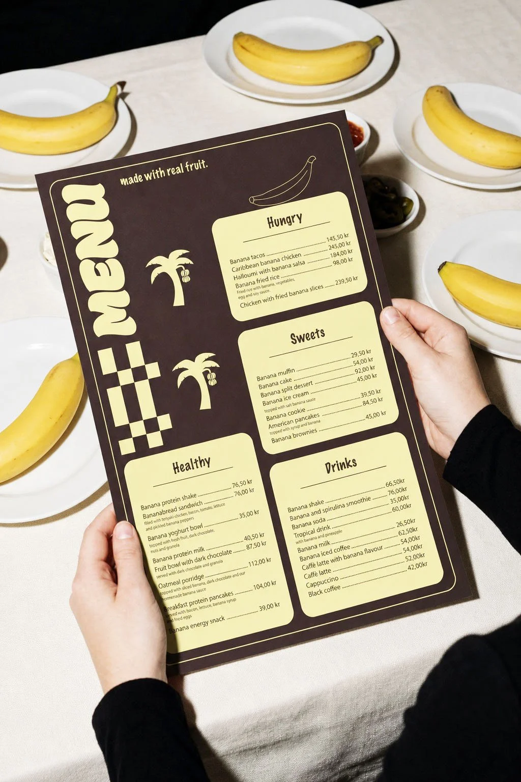 Person holding a menu with banana-themed dishes and desserts, with bananas on white plates on a table in the background.
