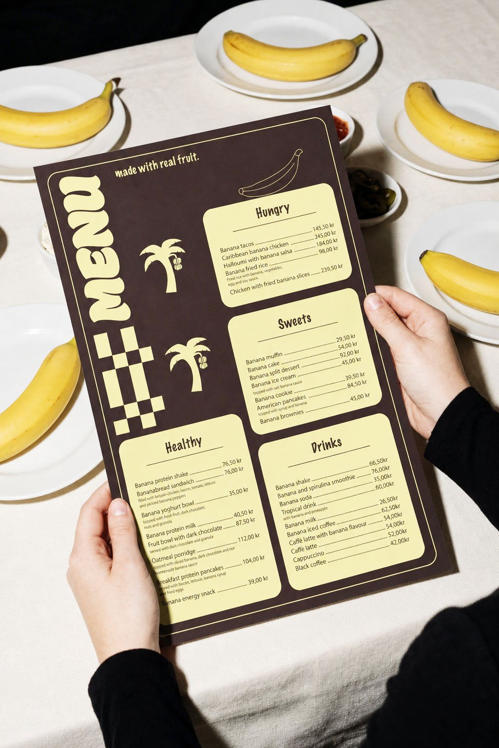 Menu card with banana-themed sections held over a table with several bananas on white plates and small bowls of condiments.