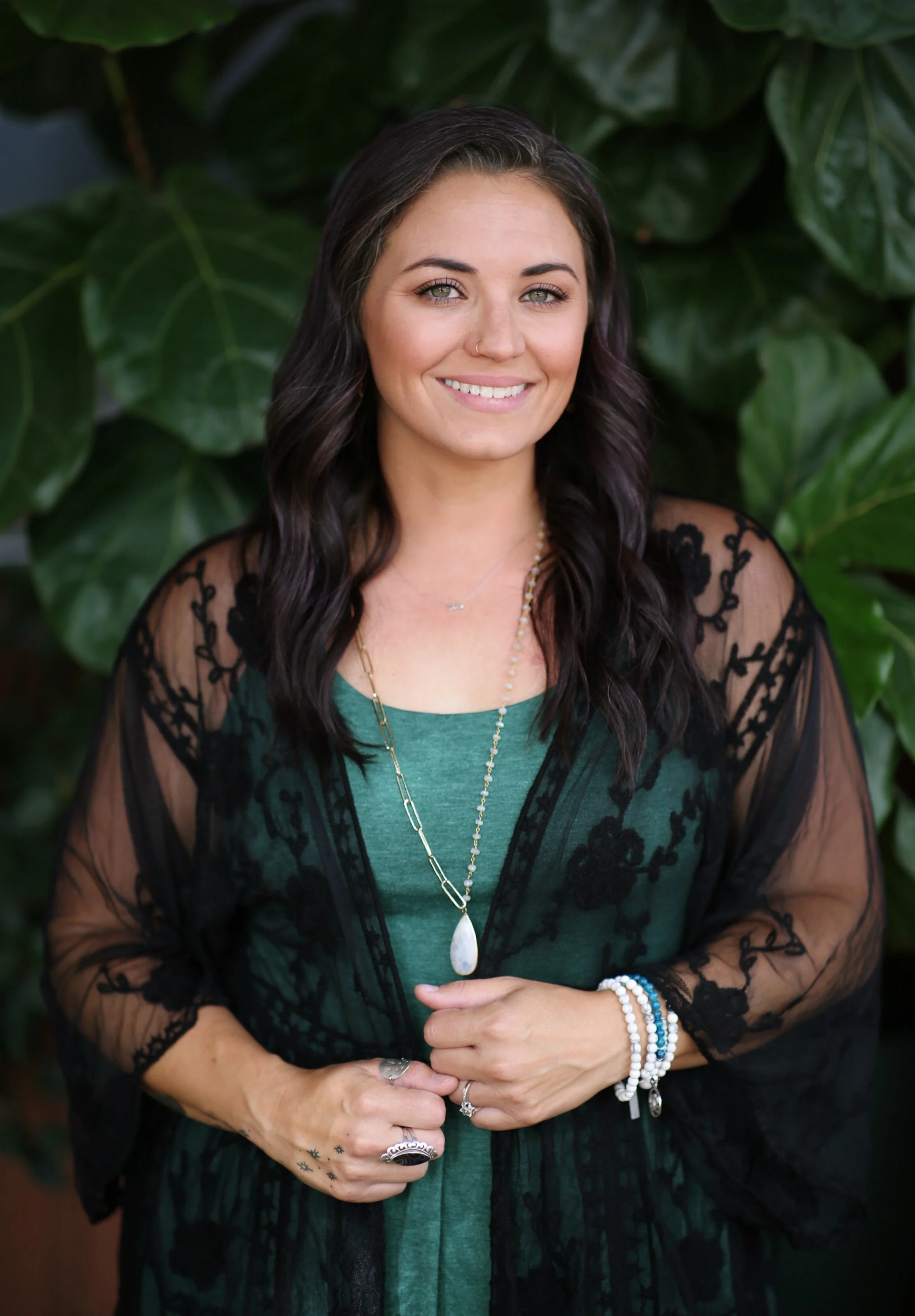 A young woman with dark wavy hair, wearing a green top and a sheer black lace embroidered cardigan, standing in front of large green leaves.