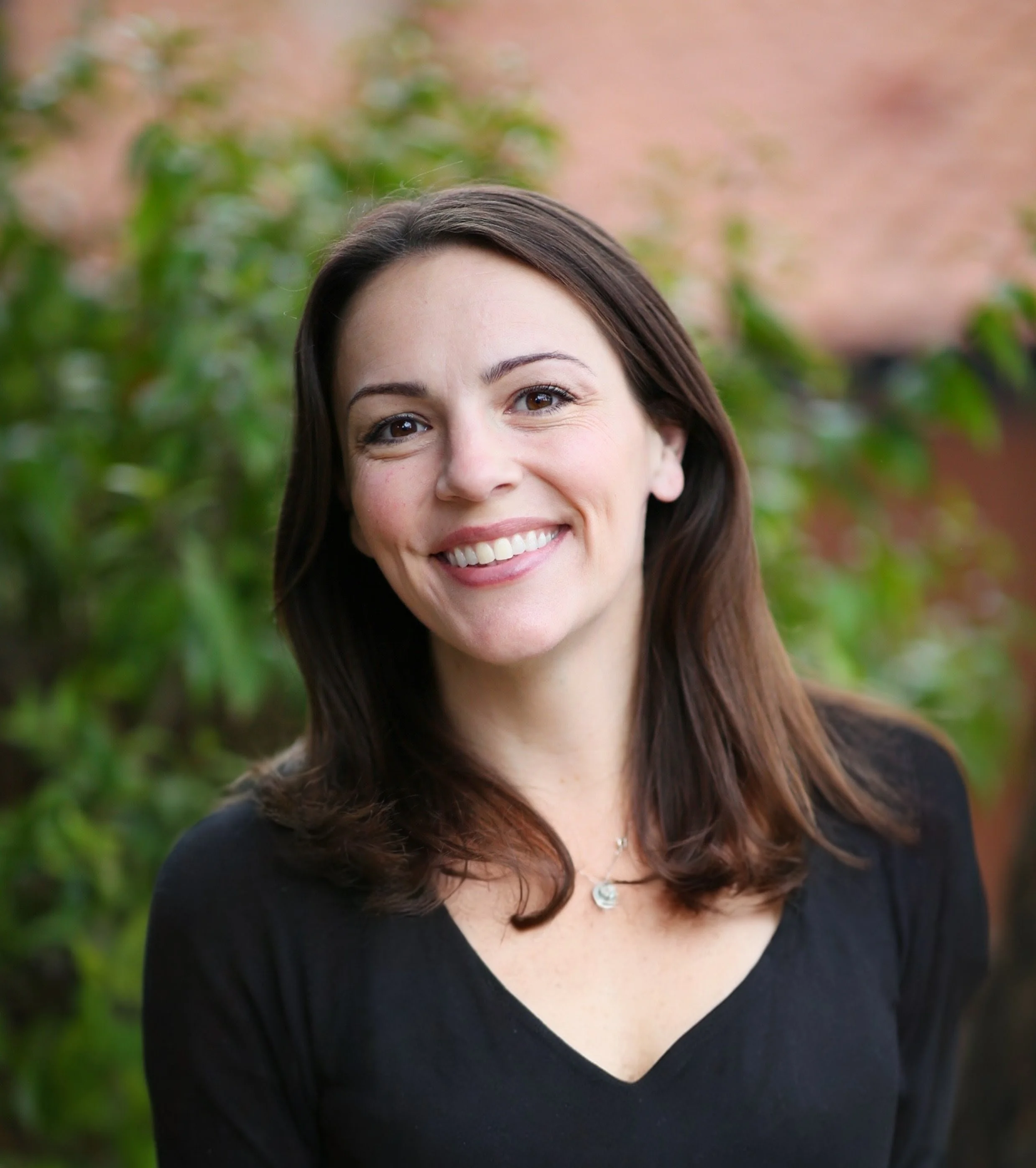 A woman with shoulder-length brown hair, smiling, wearing a black top and a silver necklace, outdoors with green foliage and a brick building in the background.
