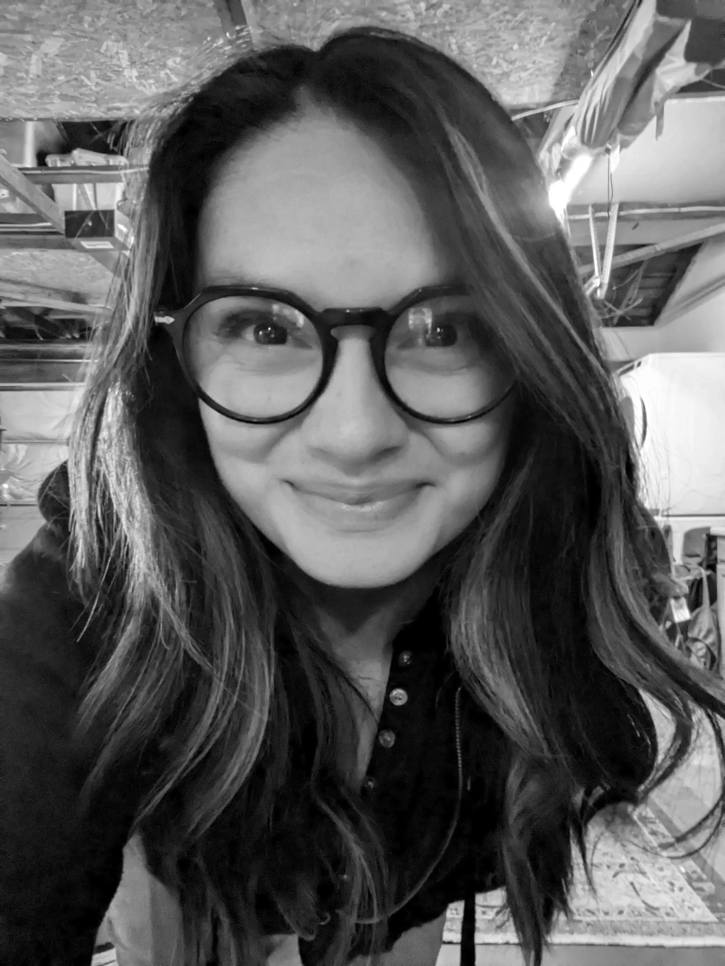 A smiling woman with long dark hair and large round glasses in a black and white photo, indoors in what appears to be a garage or workshop.