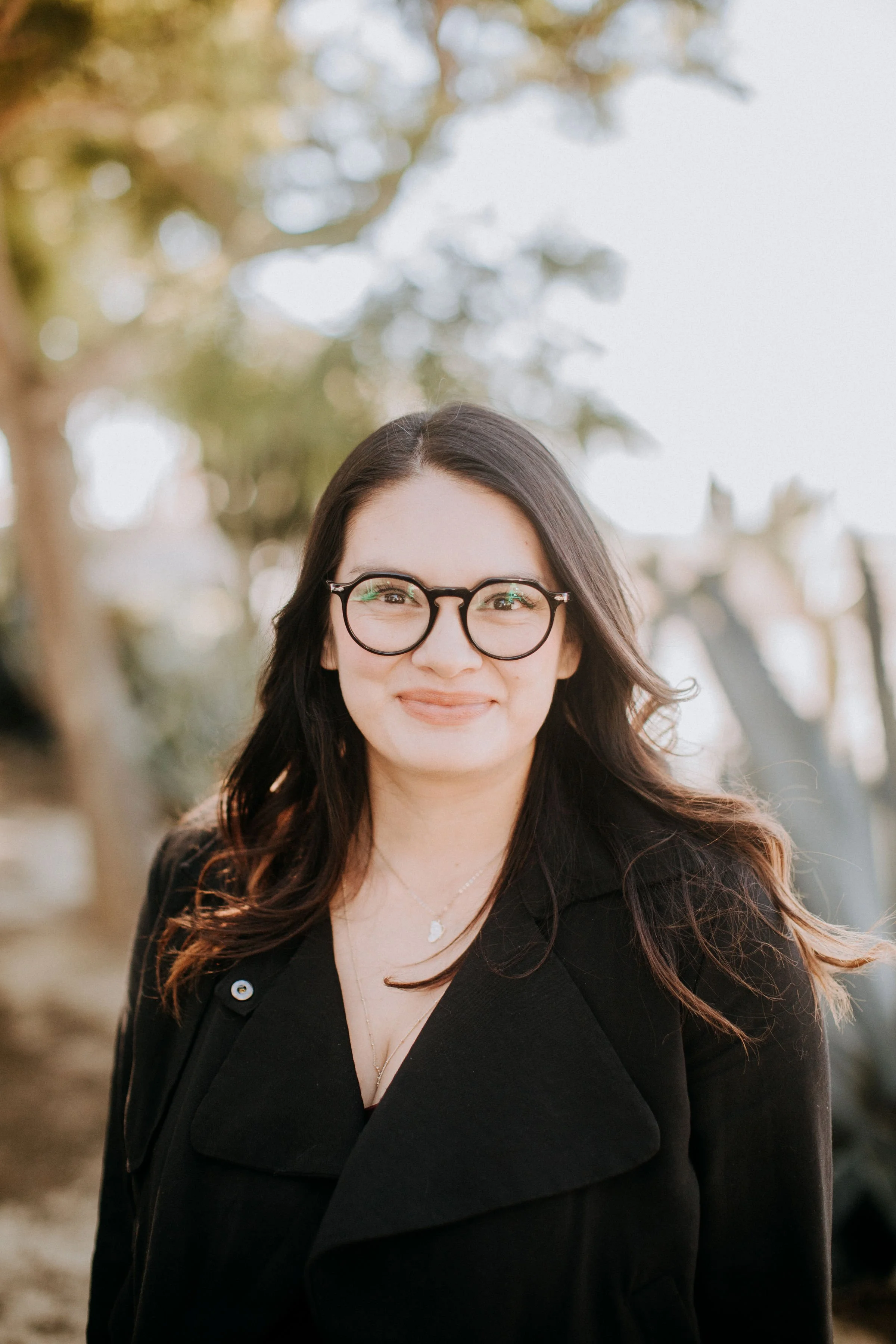 A woman with glasses and long dark hair smiling outdoors, wearing a black coat and jewelry, with trees and a fence in the background.