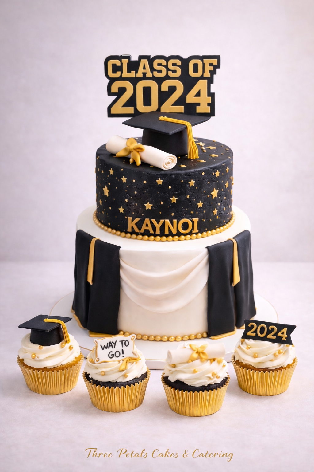 Black and white graduation cake with gold accents, topped with a graduation cap, a diploma, and a cake topper reading 'Class of 2024'. The cake has stars and the name 'Kaynoi'. Four cupcakes with graduation cap and diploma toppers are placed in front.